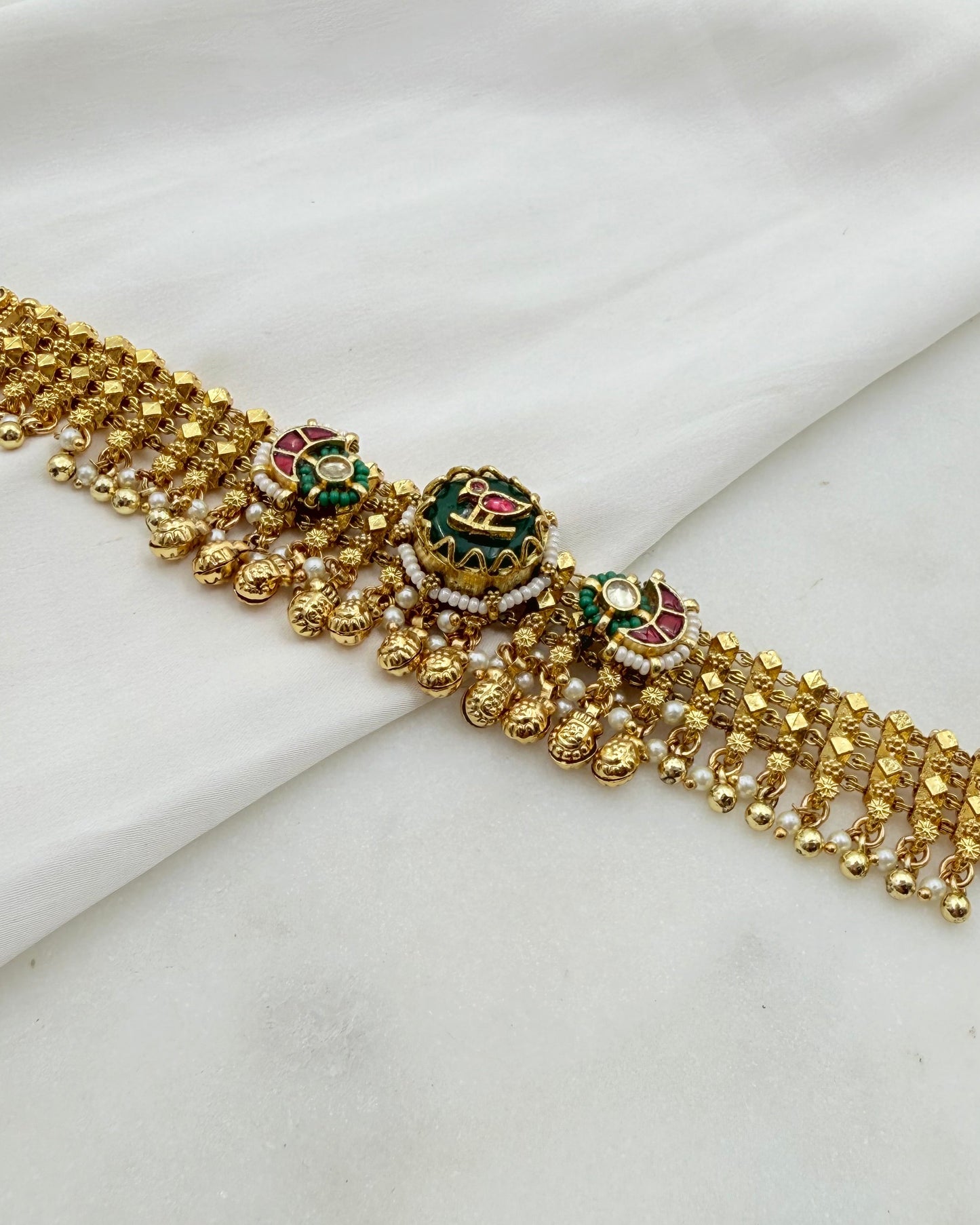 A gold-plated traditional Indian choker necklace with intricate beadwork, floral motifs, and green and pink enamel accents, displayed on a draped white fabric background.