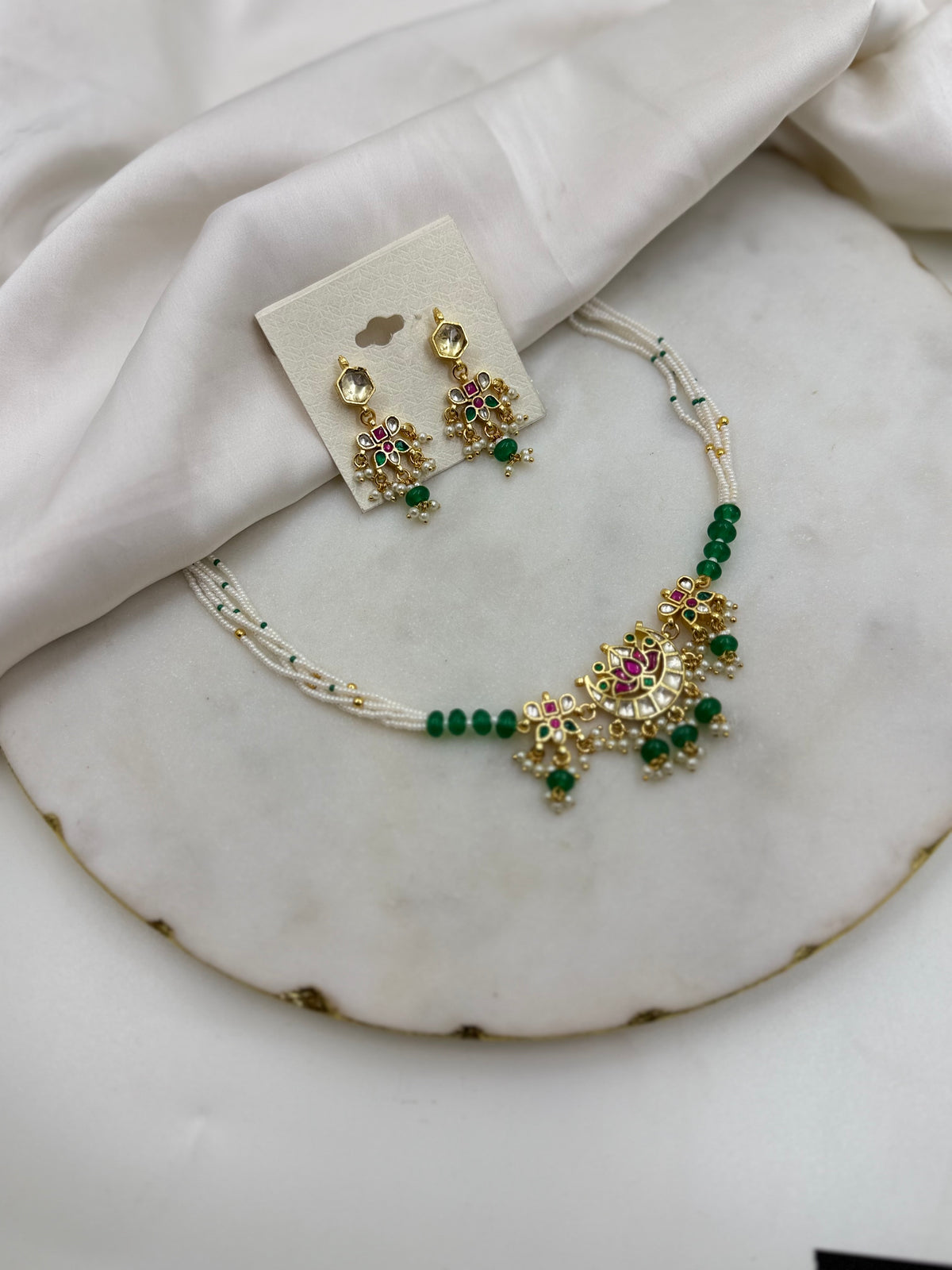 Gold-toned traditional Indian jewelry set featuring a necklace and matching earrings with green beads, pink stones, and gold floral motifs, displayed on a white marble slab with white fabric.