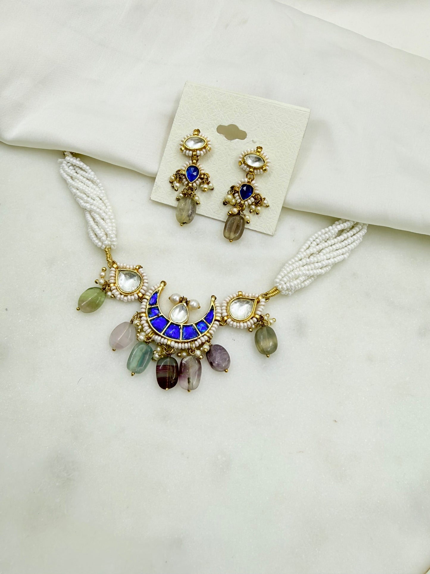 A gold-toned jewelry set featuring a necklace and matching earrings displayed on a white fabric surface. The necklace has white braided cords, a blue crescent pendant with gold detailing, and dangling multicolored teardrop beads.