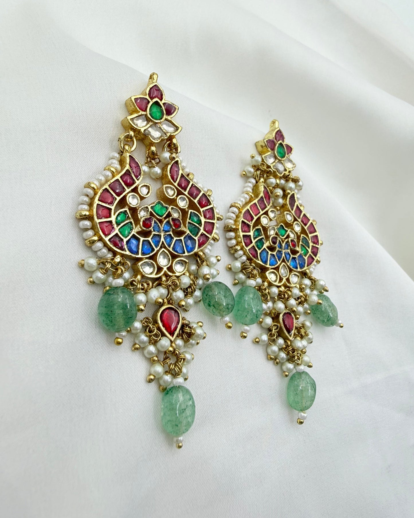 A pair of ornate gold-toned chandelier earrings decorated with multicolored enamel, white faux pearls, and green gemstone drops, displayed on a white fabric background.