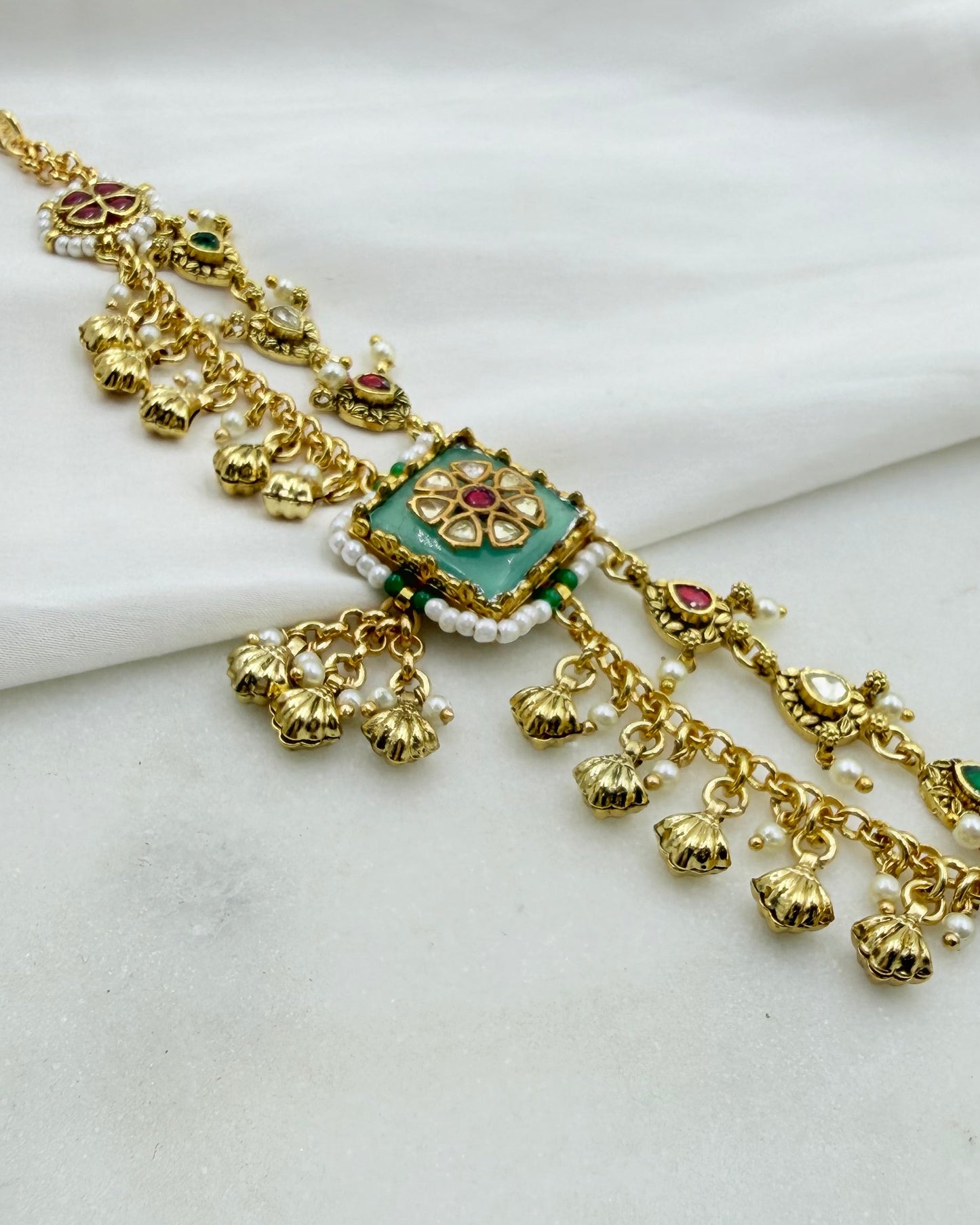 Gold-toned traditional Indian jewelry piece with a central turquoise green square, red and green stone accents, and dangling bell charms, displayed on a white fabric background.