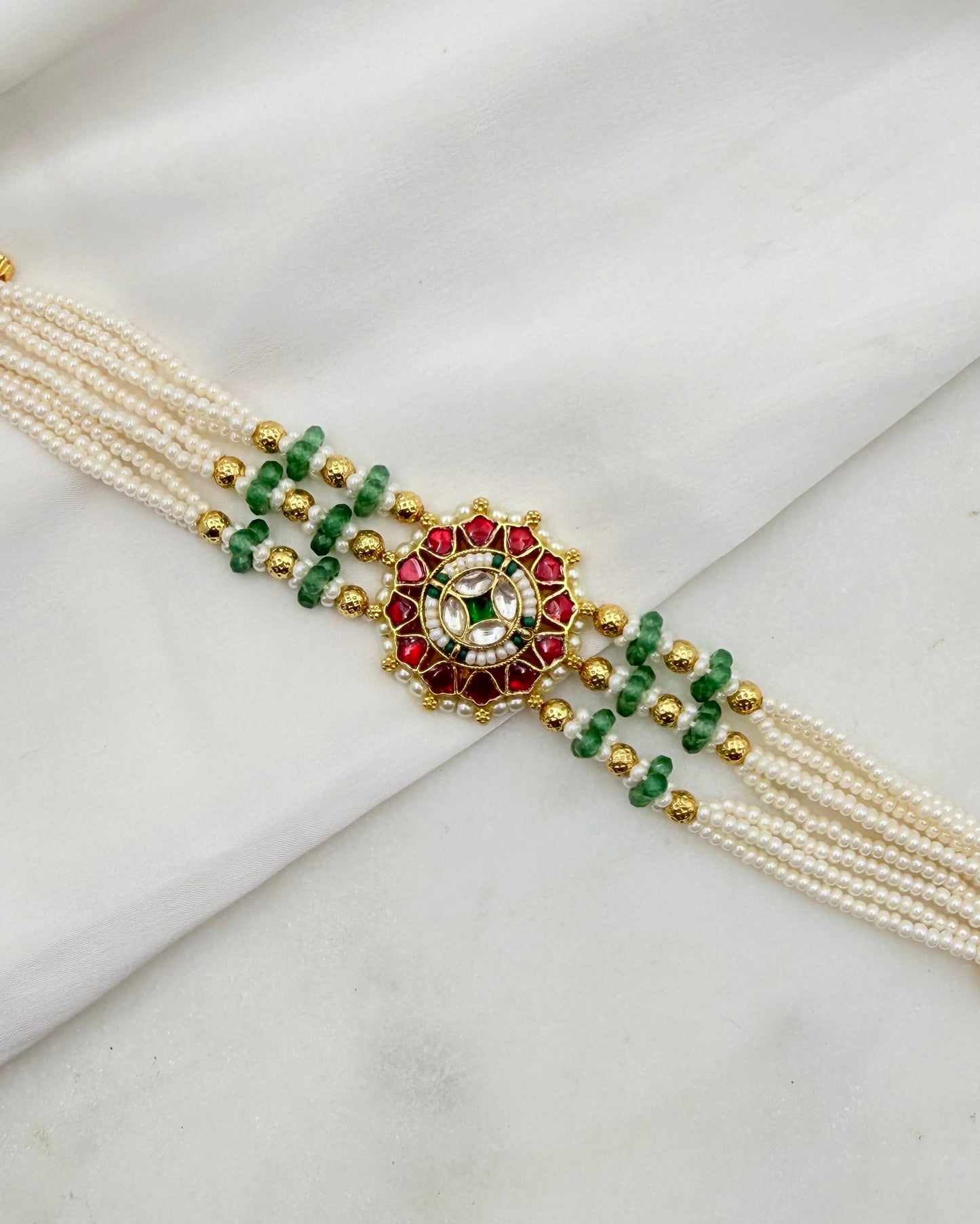 A traditional Indian pearl bracelet (Kara) with gold-plated metal, featuring cream-colored pearls, green and red stones, and gold bead accents, laid flat on a white draped fabric background.