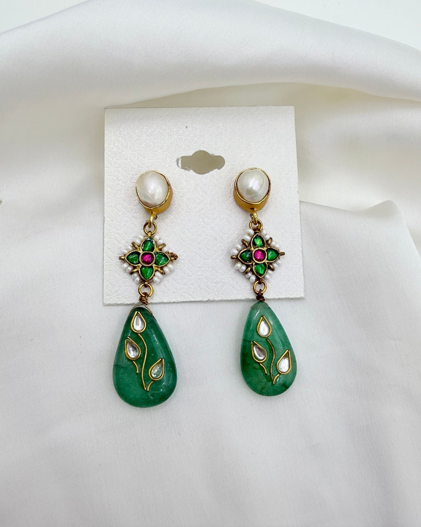 A pair of traditional Indian gold-plated dangle earrings with green stone teardrop pendants, decorated with white stones and pink/red inlays, displayed on a white card against a draped white fabric background.