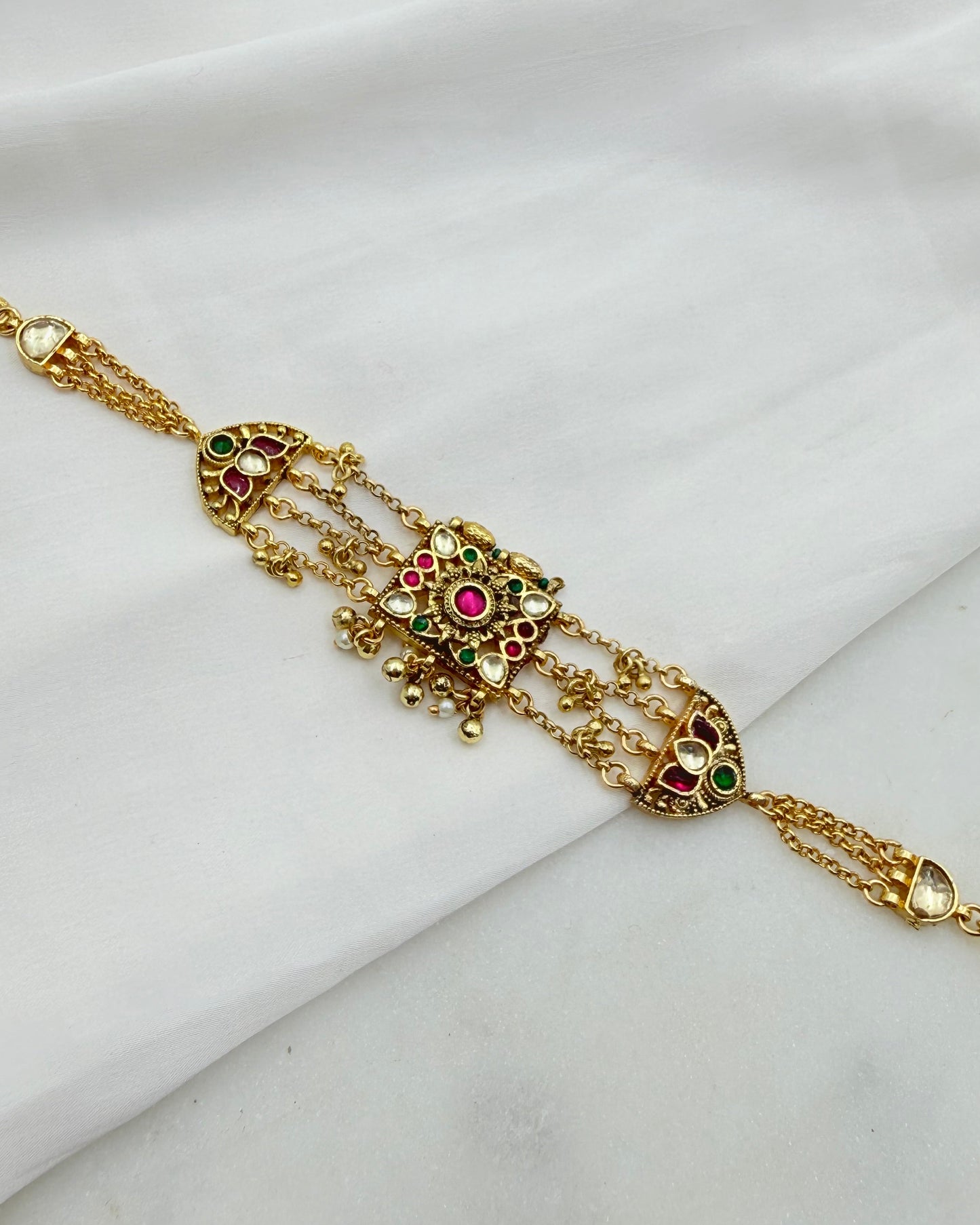 Gold-toned traditional Indian jewelry piece with intricate filigree, small dangling beads, and colorful stone accents in red, green, and pink, displayed on a draped white fabric background.