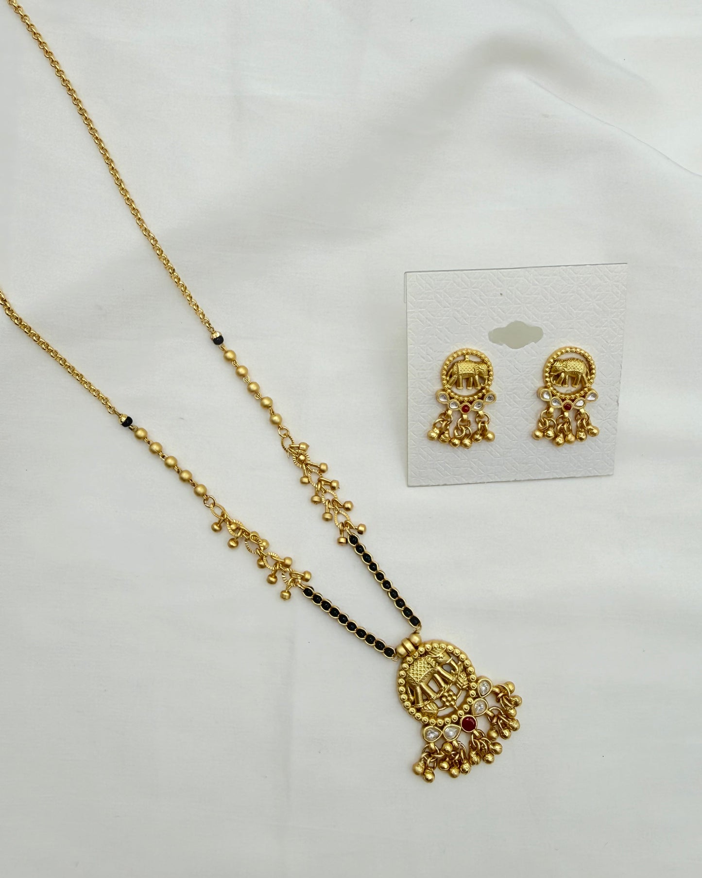Gold-toned traditional jewelry set featuring a necklace with a detailed pendant and matching stud earrings, displayed on a white fabric background next to a white card.