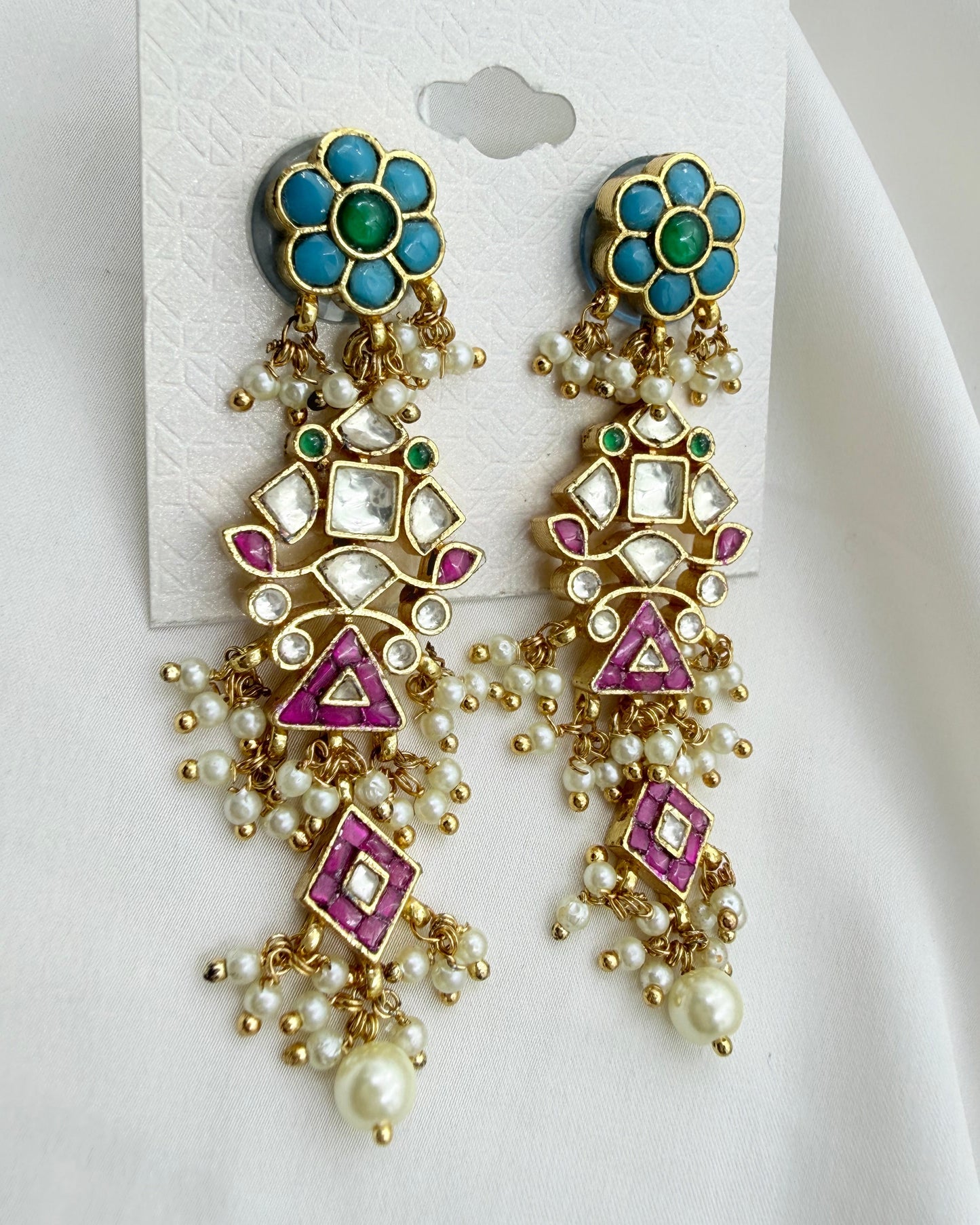A pair of ornate gold-toned statement earrings with turquoise blue flower-shaped studs, green accents, white faux pearls, and pink enamel geometric details, displayed on a white card against a white fabric background.