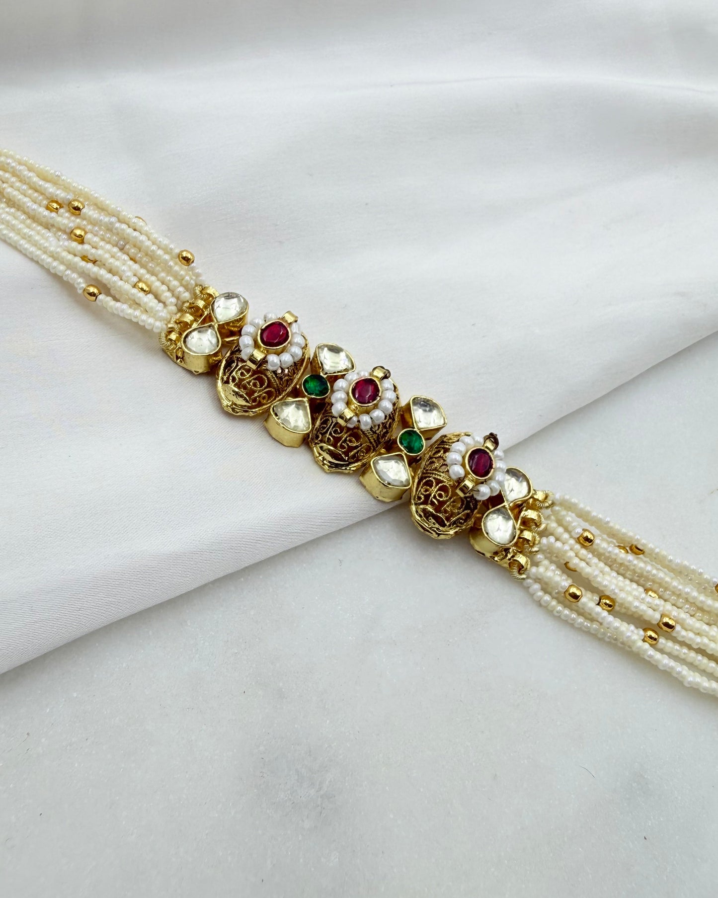 A traditional Indian gold-plated choker necklace with multiple strands of white pearl beads and decorative floral motifs set with white stones, red rubies, and green emeralds, displayed on a draped white fabric background.