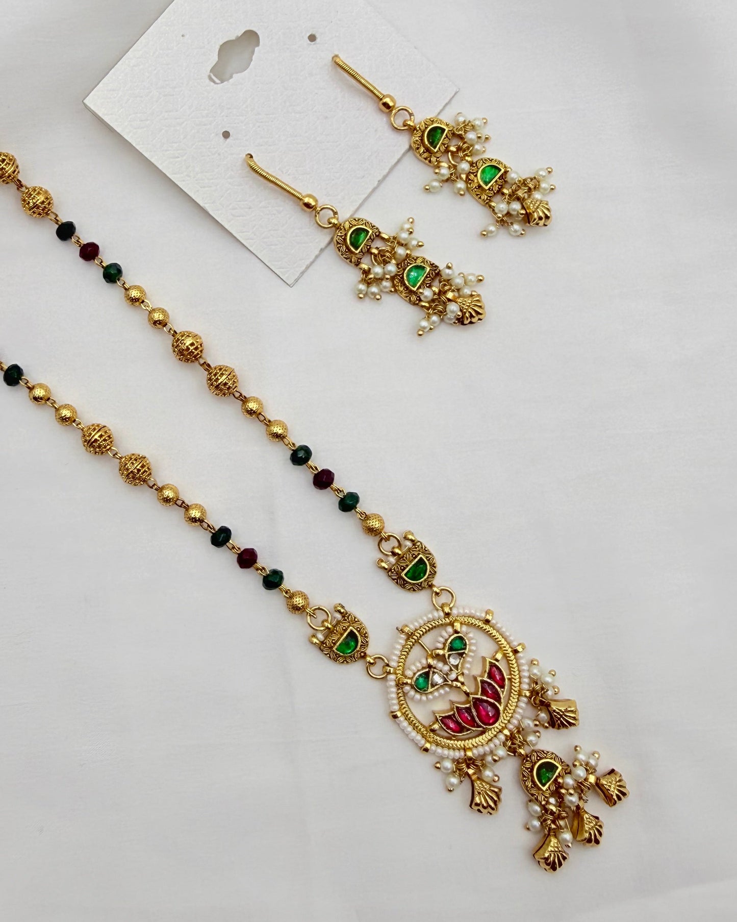 Gold-toned traditional jewelry set featuring a beaded necklace with a detailed peacock pendant, matching drop earrings, and a small pearl card, displayed on a white fabric background.