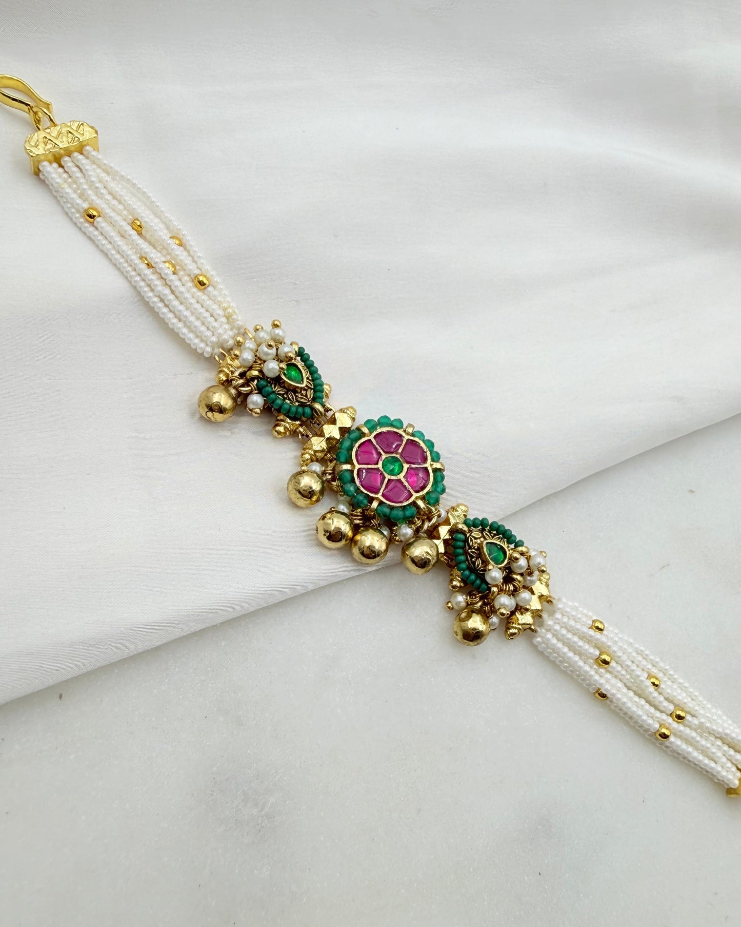 A traditional Indian multi-strand pearl bracelet with gold-plated metal details, featuring a central floral motif in pink and green enamel, displayed on a white fabric background.