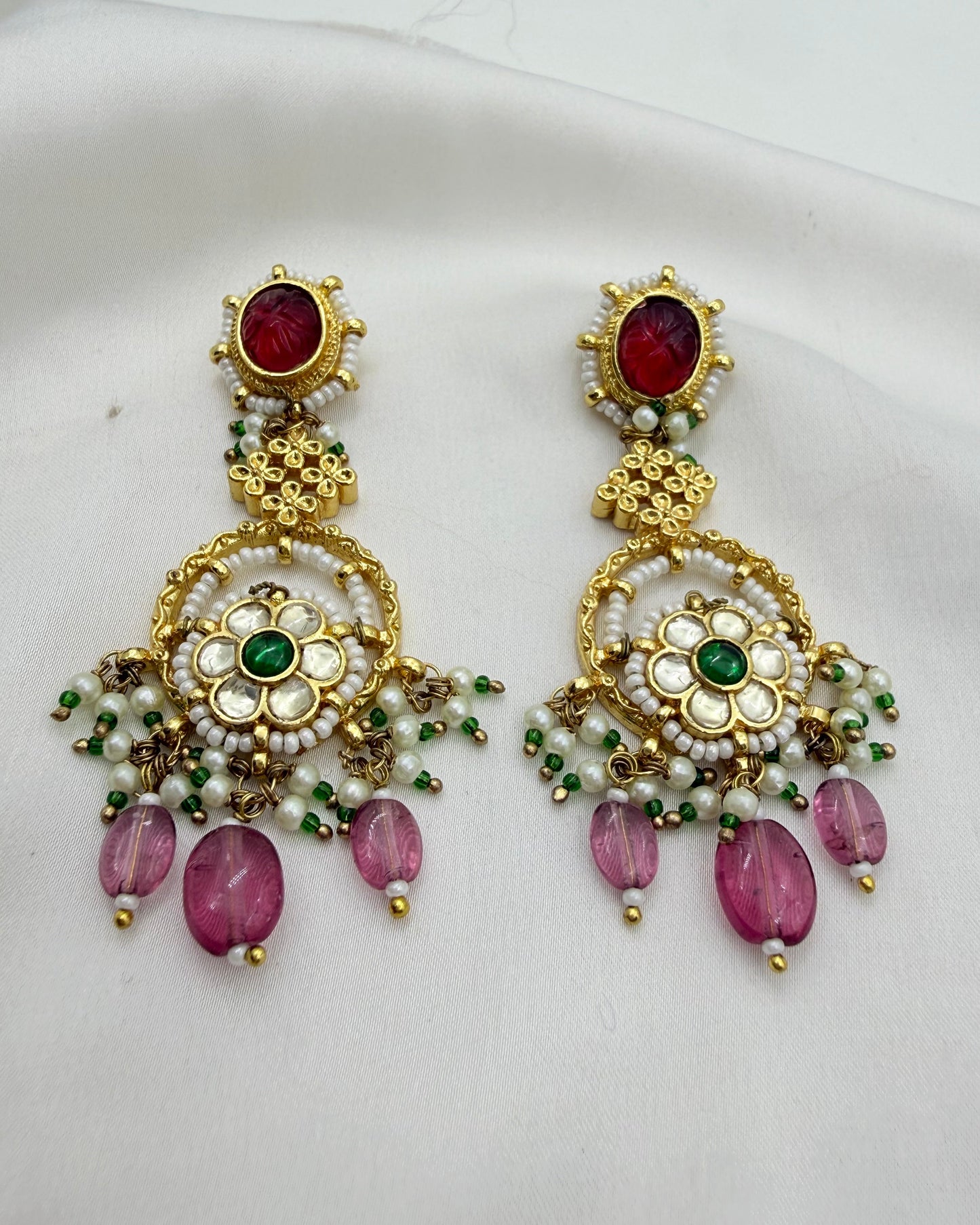A pair of ornate gold-plated Indian chandelier earrings decorated with red cabochon stones, white faux pearls, green beads, and pink teardrop beads, displayed on a white fabric background.