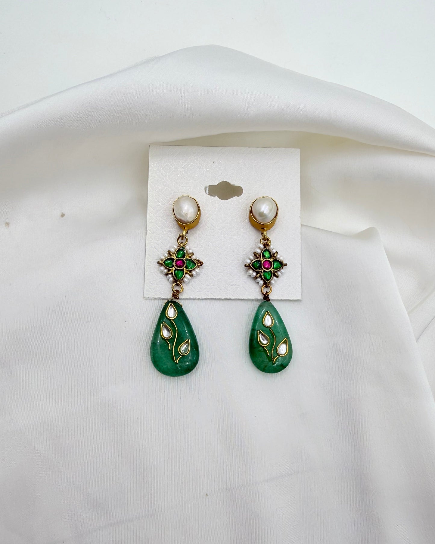 A pair of gold-tone drop earrings with white pearl studs, green floral charms with red and green stones, and green teardrop charms decorated with gold and white leaf details, displayed on a white card over a draped white fabric background.