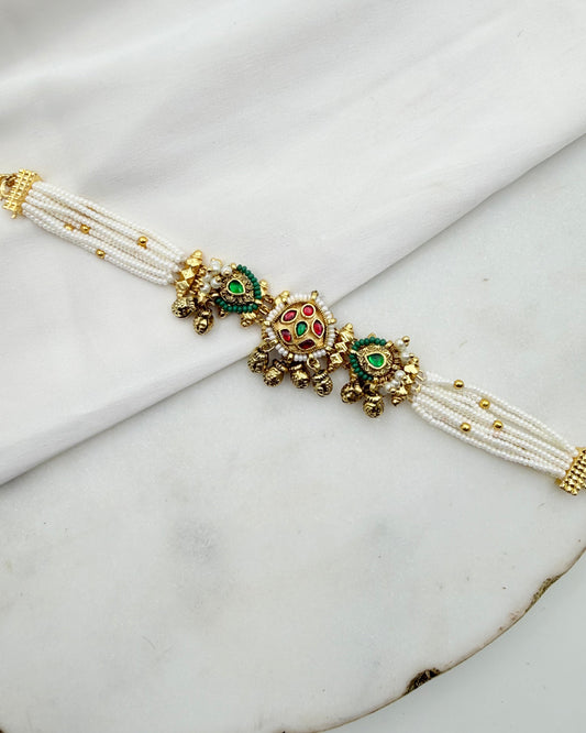 A traditional Indian multi-strand pearl bracelet with gold-plated borders and ornate central motifs featuring green, red, and gold enamel details, displayed on a white fabric and marble surface.