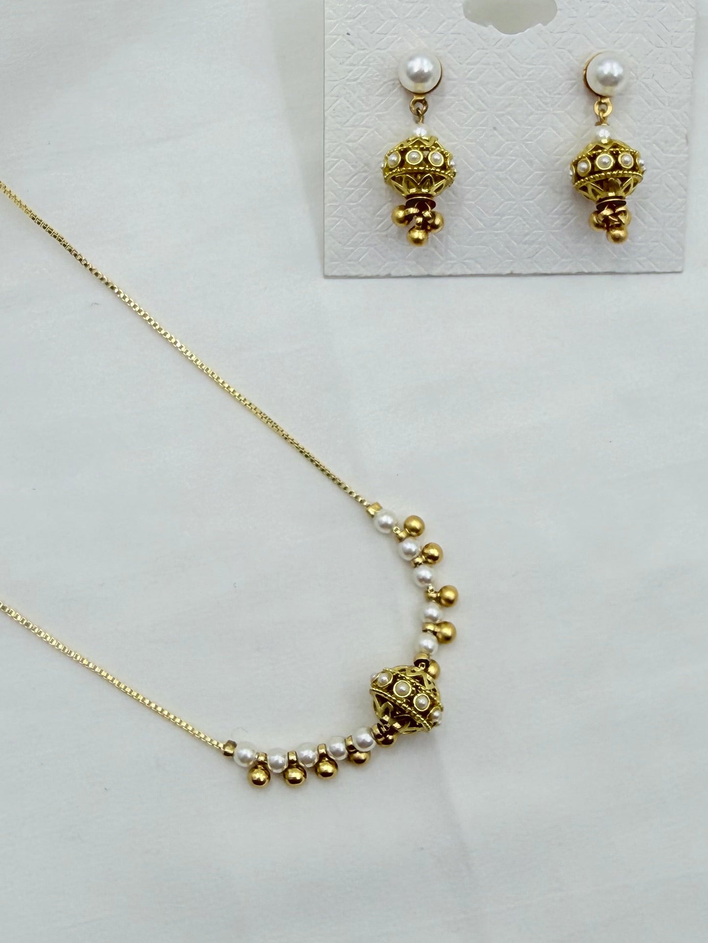 Gold necklace and matching drop earrings with small white pearls, displayed on a white card over a white fabric background.