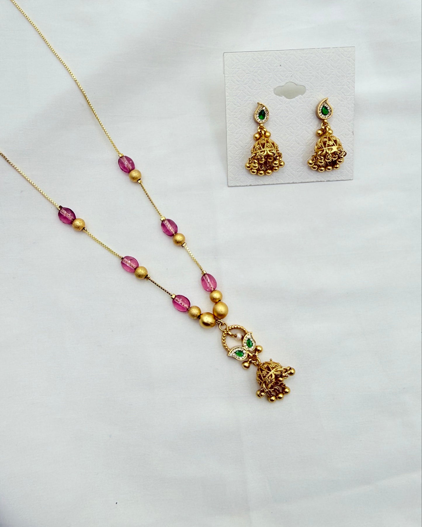 Gold-toned jewelry set on a white fabric background, including a necklace with pink and gold beads and a dangling elephant motif, plus matching earrings displayed on a small card.