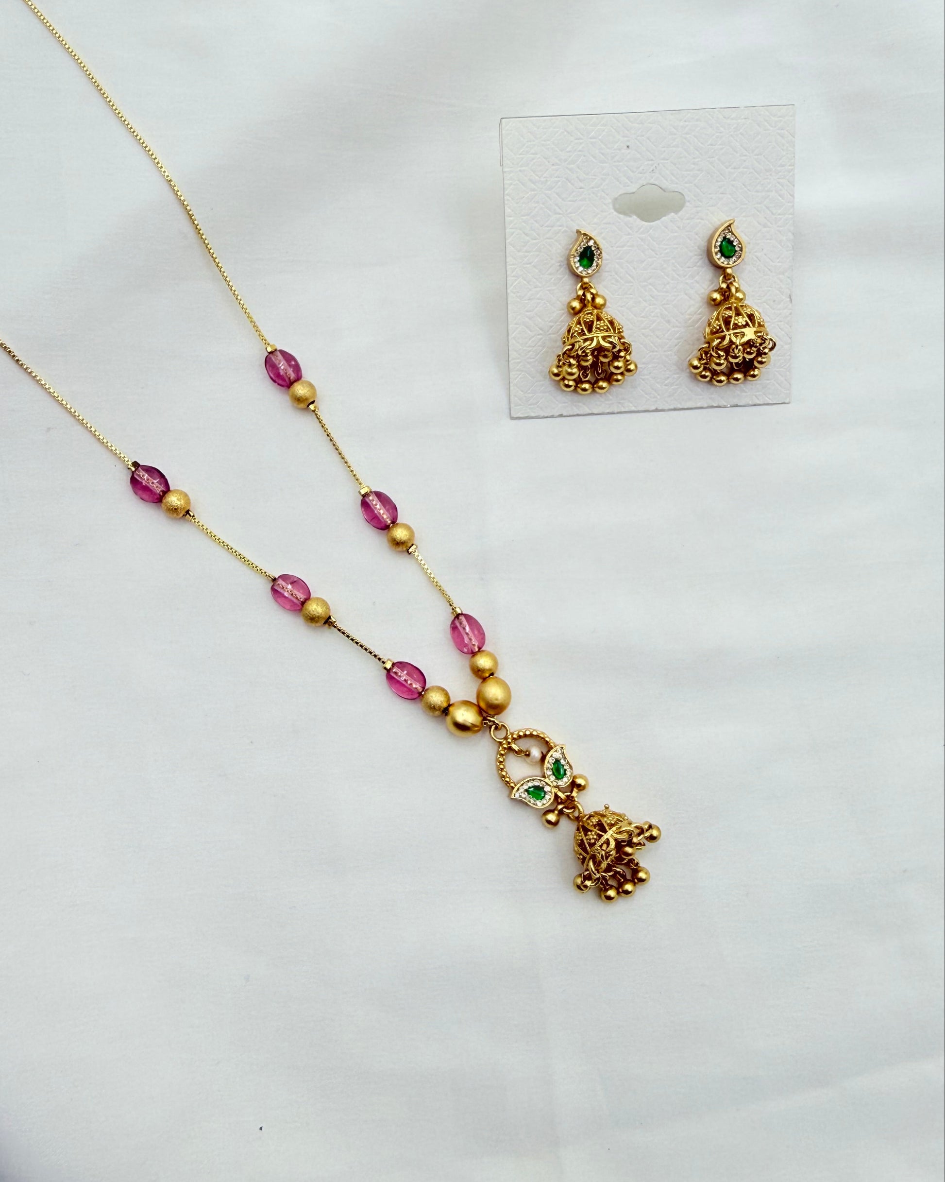 Gold-toned jewelry set on a white fabric background, including a necklace with pink and gold beads and a dangling elephant motif, plus matching earrings displayed on a small card.