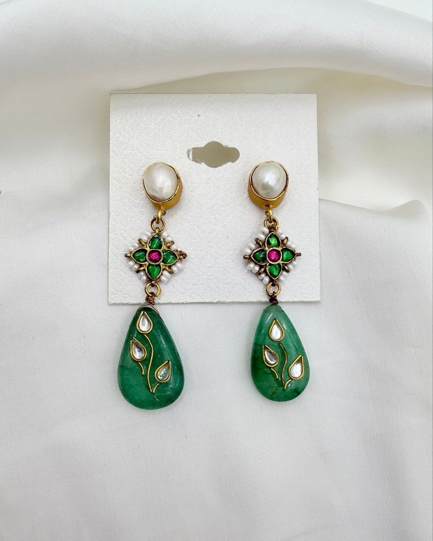 A pair of gold-plated dangle earrings with white pearl studs, green enamel starburst details with red and pink accents, and green teardrop pendants decorated with gold and white enamel leaf motifs, displayed on a white card against a white fabric background.