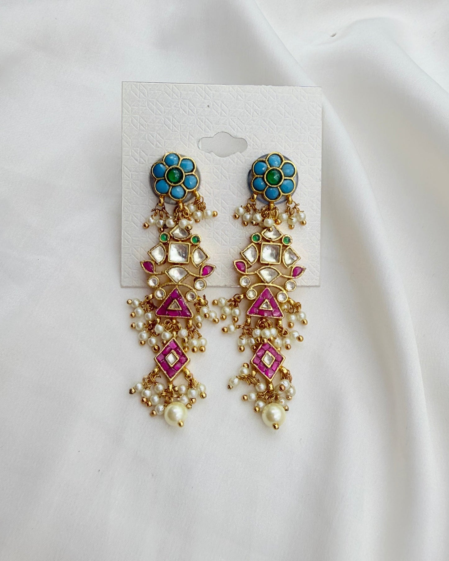 A pair of ornate gold-toned dangle earrings with turquoise, green, pink, and white stone accents, displayed on a white card against a white fabric background.