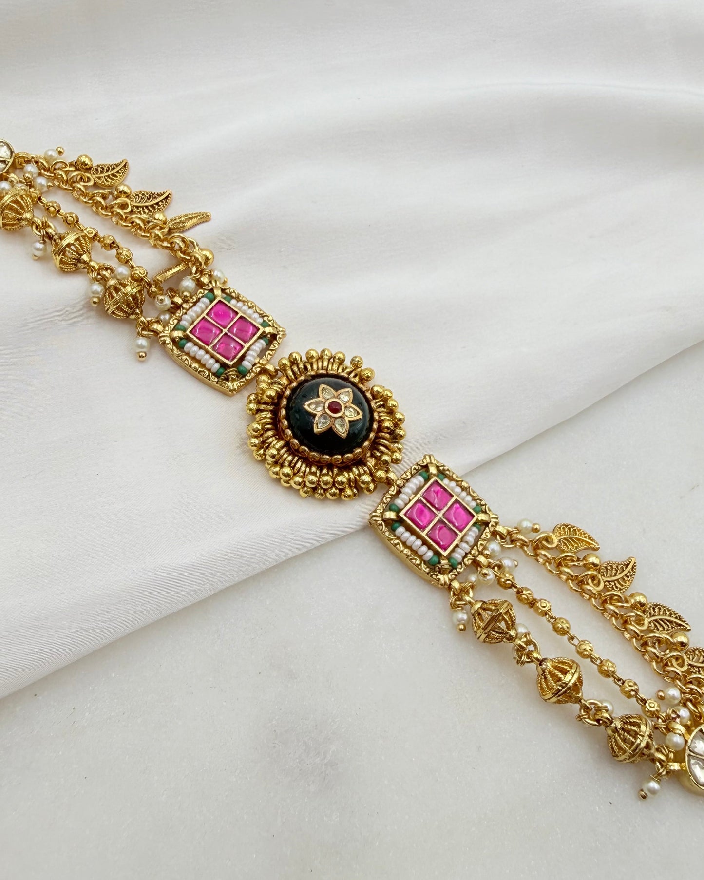 An ornate gold-toned traditional Indian waist chain or belt featuring a central black oval medallion with a floral motif, flanked by two square panels with pink and green accents, set against a draped white fabric background.