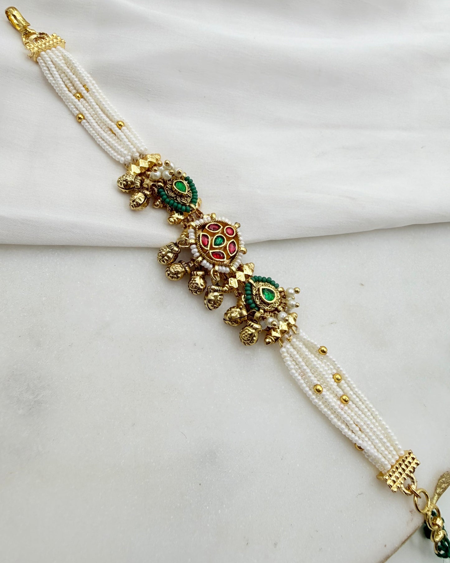A traditional Indian multi-strand pearl bracelet with gold-plated filigree and green and red enamel details, displayed on a white fabric background.