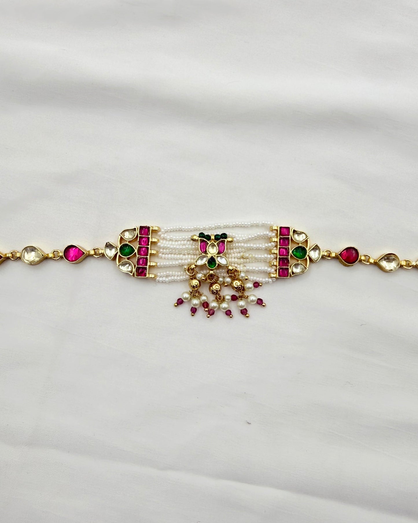 A gold-plated traditional Indian bracelet (lalka) featuring white pearl strands, green and pink stones, and a central gold-toned decorative element, displayed on a white fabric background.