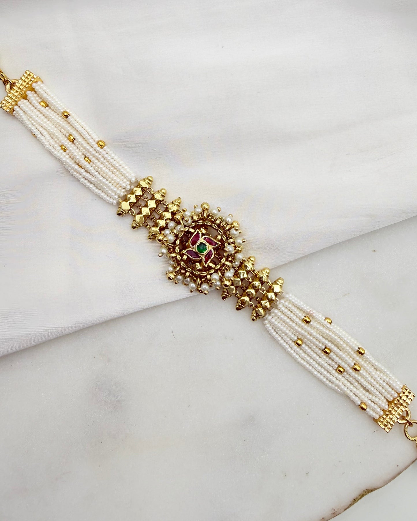 A traditional Indian multi-strand pearl bracelet with gold-plated metal beadwork and a central red and green stone motif, displayed on a white marble surface.