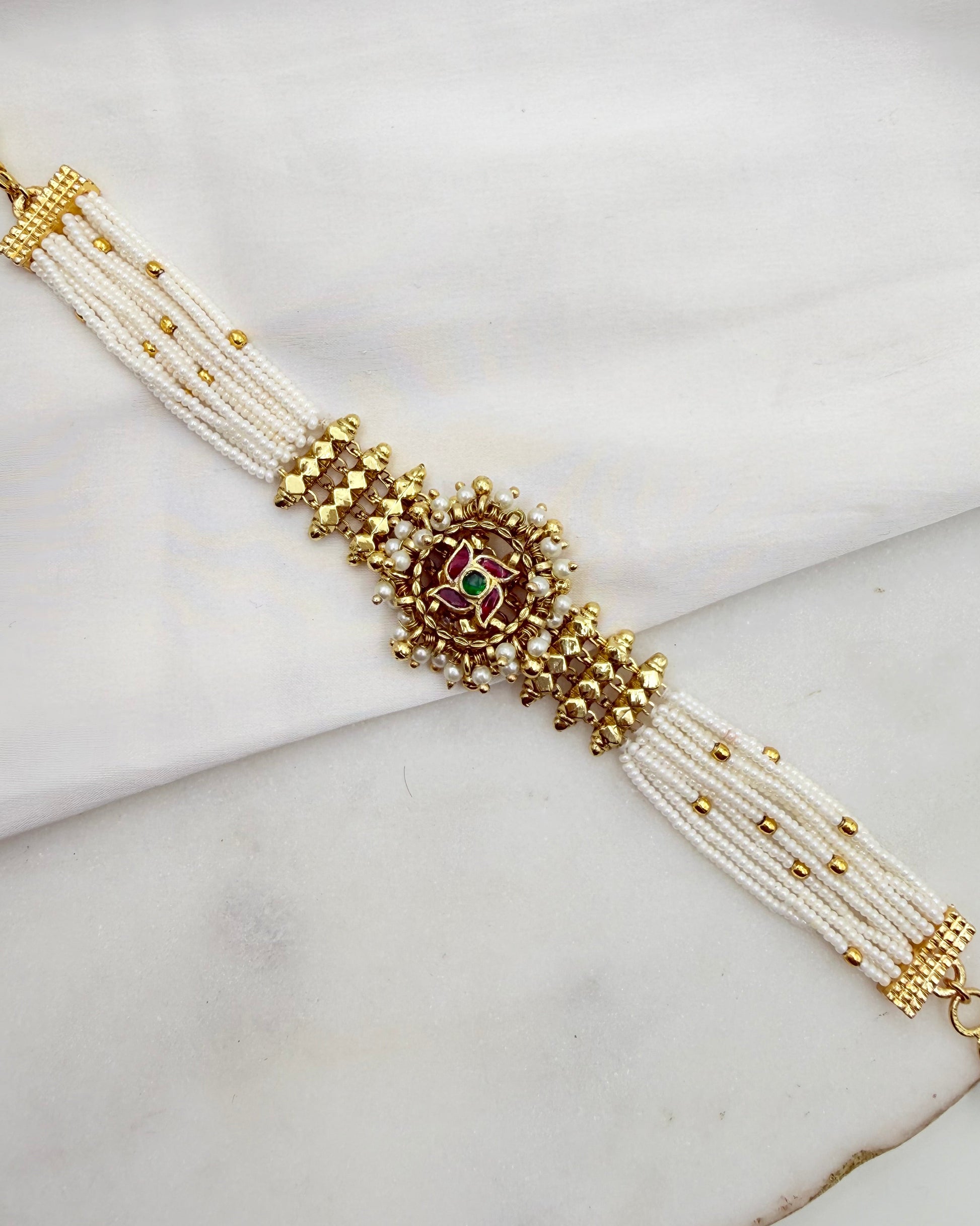 A traditional Indian multi-strand pearl bracelet with gold-plated metal beadwork and a central red and green stone motif, displayed on a white marble surface.