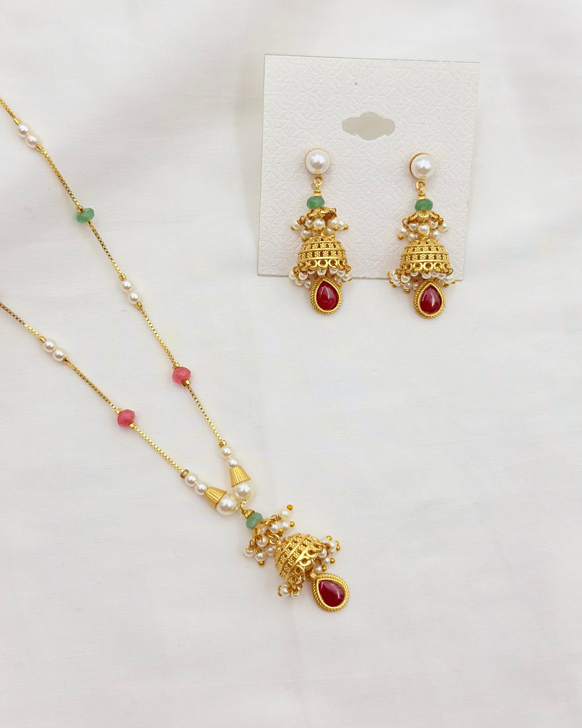 Gold-toned jewelry set featuring a necklace and matching earrings with pearl-like white beads, small green accents, and red teardrop stones, displayed on a white background with a card.