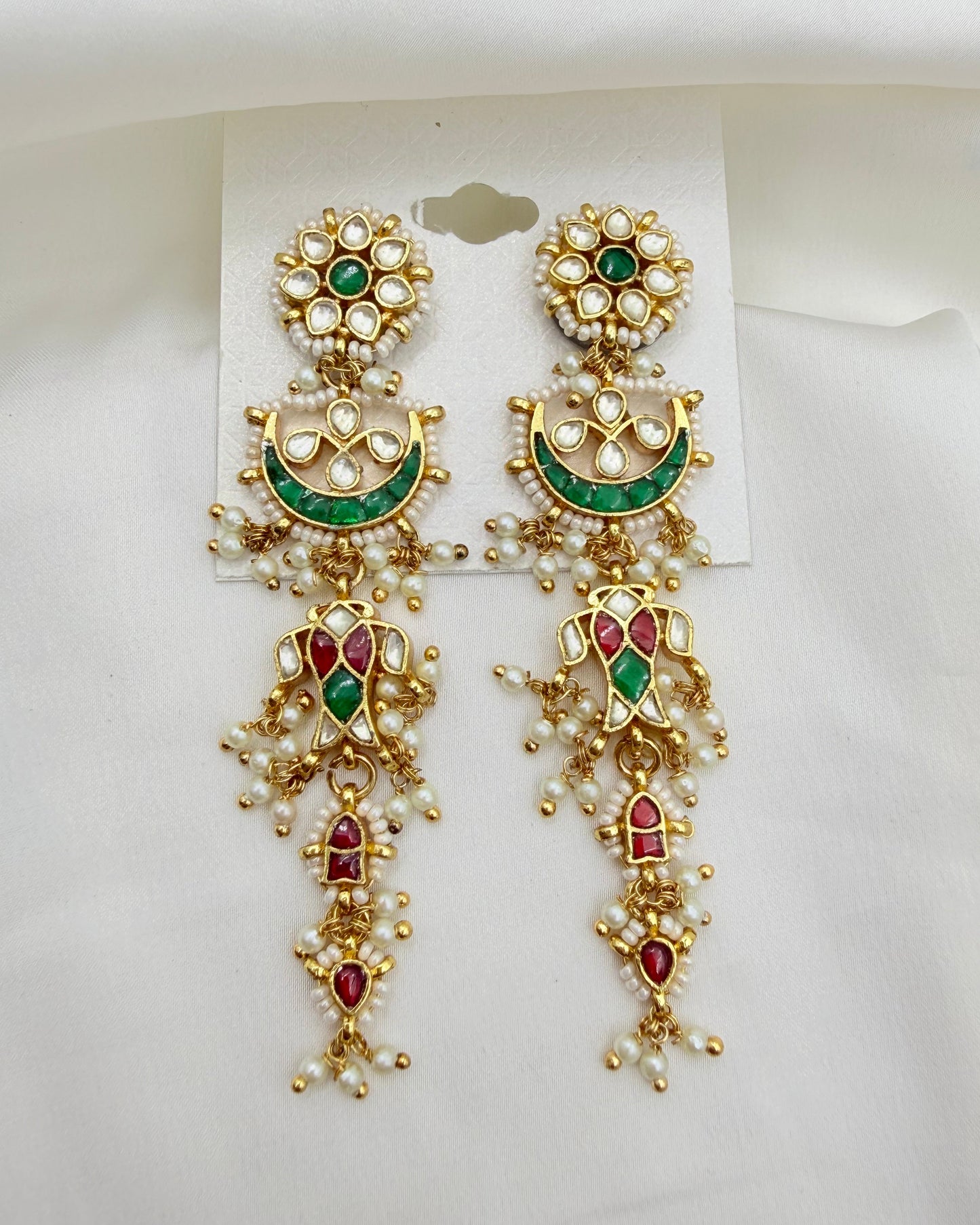 A pair of ornate gold-toned Indian Kundan dangle earrings with green and red enamel details, set with white faux pearls, displayed on a white card against a white fabric background.