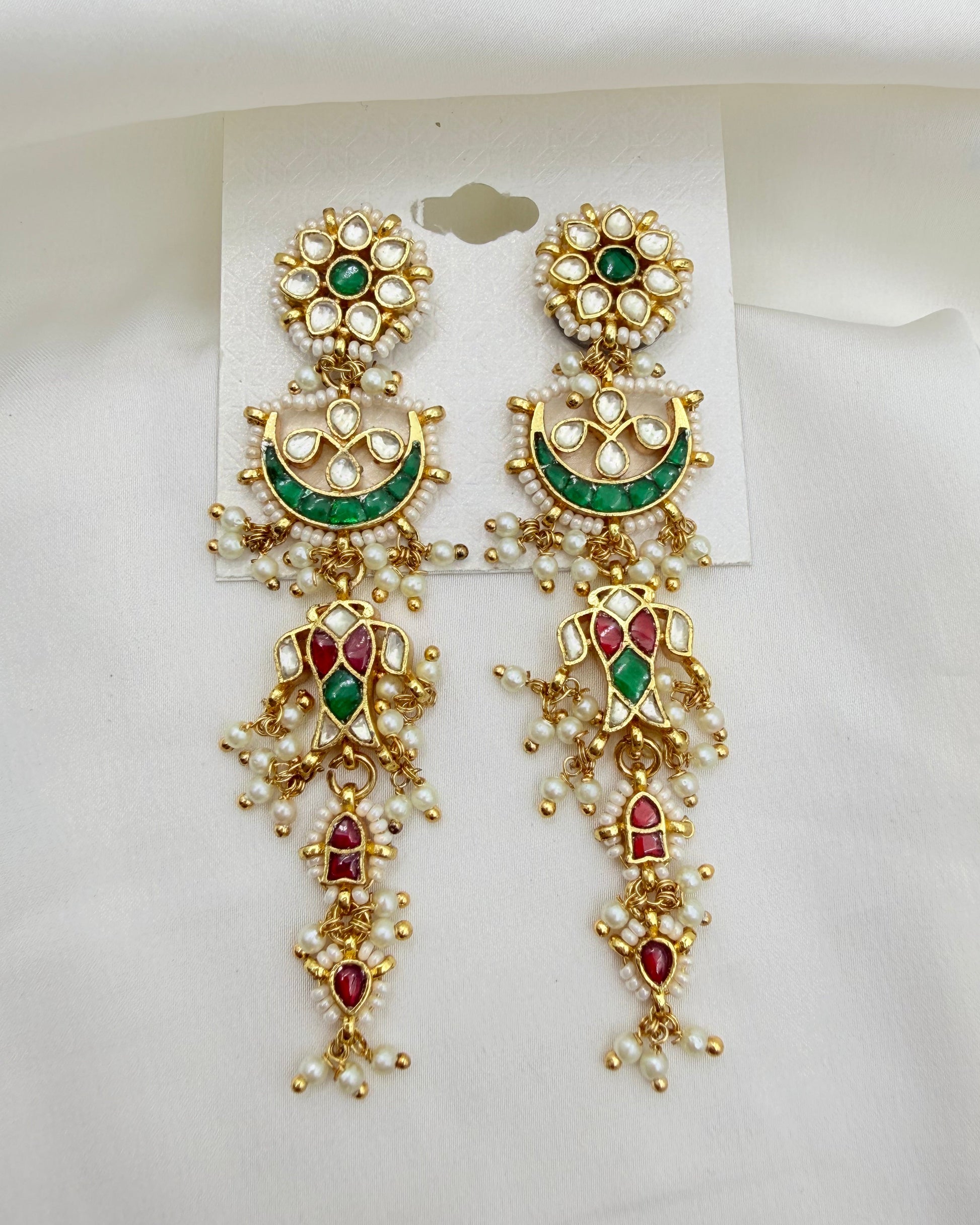 A pair of ornate gold-toned Indian Kundan dangle earrings with green and red enamel details, set with white faux pearls, displayed on a white card against a white fabric background.