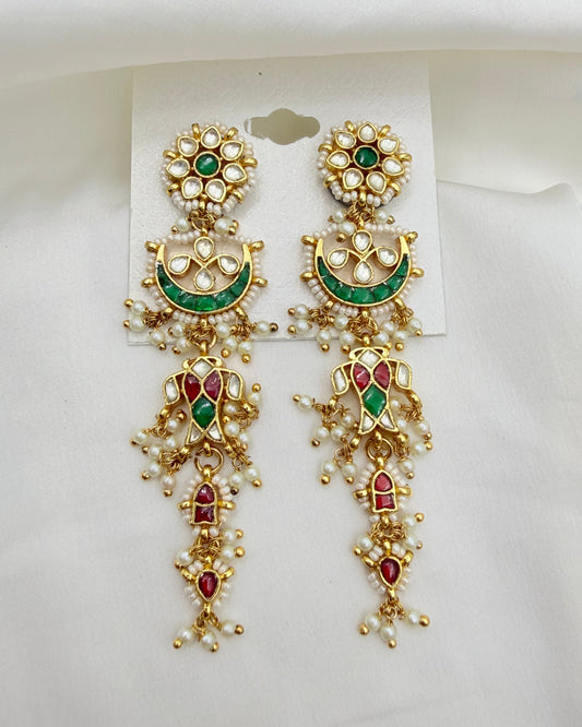 A pair of ornate gold-toned Indian Kundan dangle earrings with green and red enamel details, set with white faux pearls, displayed on a white card against a white fabric background.
