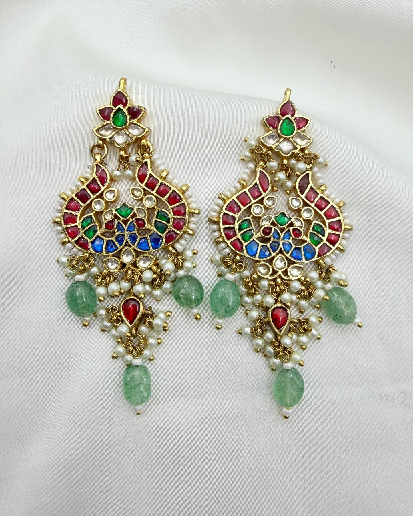 A pair of ornate gold-toned Indian-style dangle earrings decorated with red, green, and blue enamel, white faux pearls, and green teardrop beads, displayed on a white fabric background.