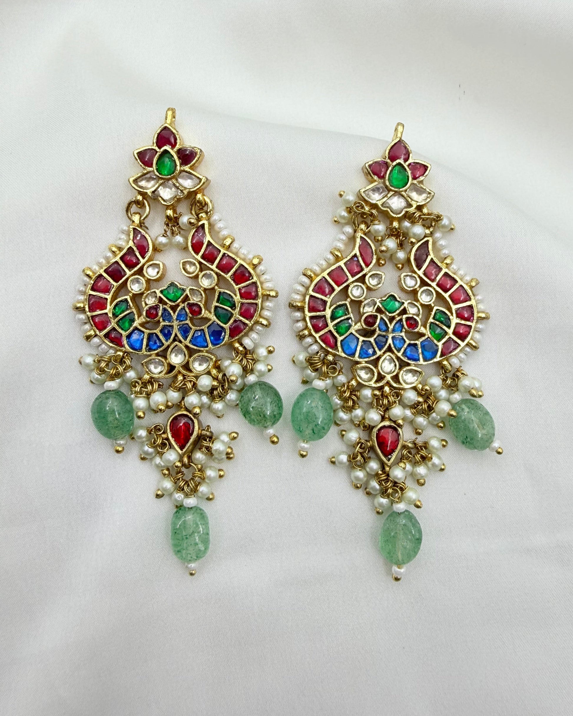 A pair of ornate gold-toned Indian-style dangle earrings decorated with red, green, and blue enamel, white faux pearls, and green teardrop beads, displayed on a white fabric background.