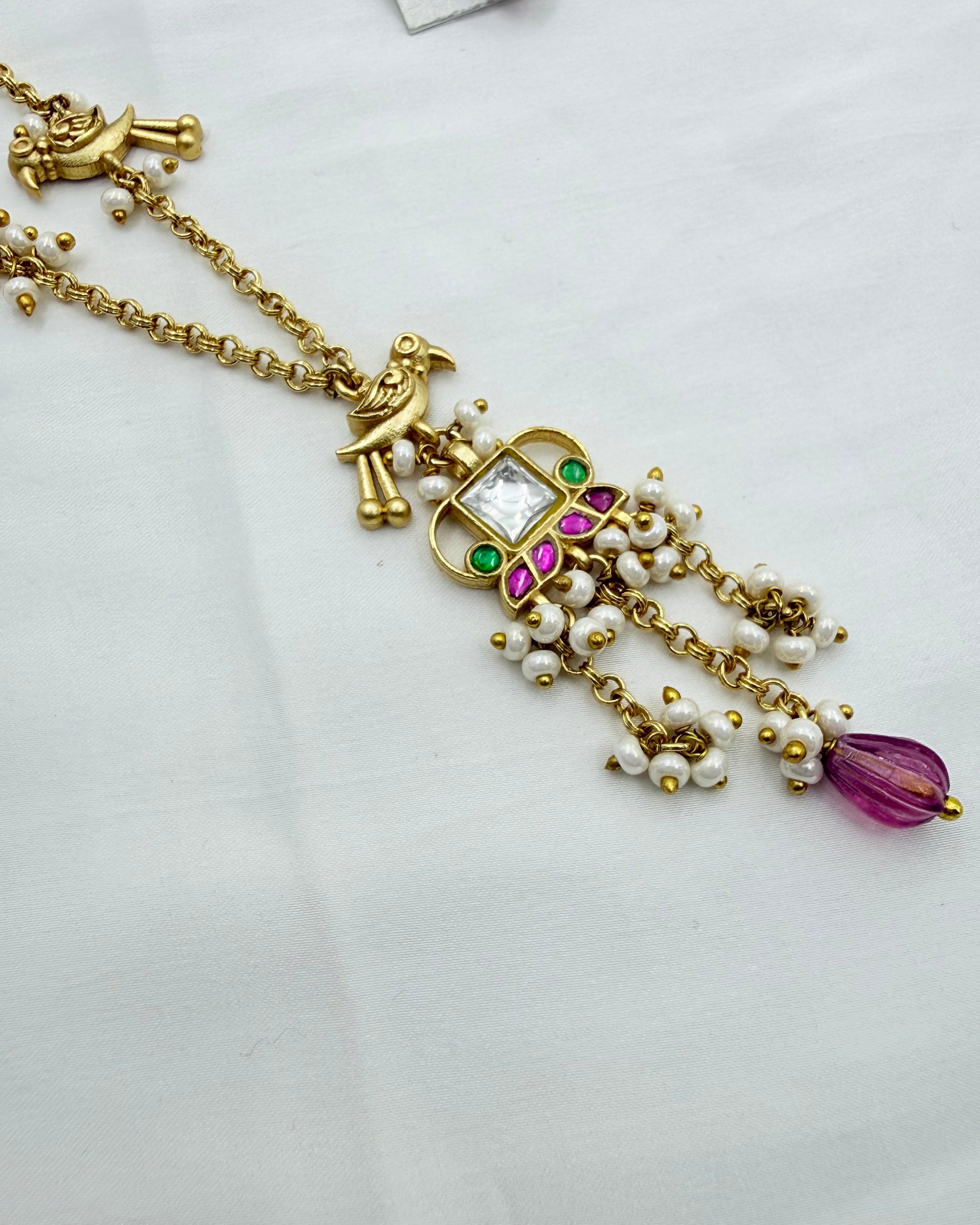 Gold-toned traditional necklace featuring a bird motif, a central square pendant with a clear stone and green and pink accents, dangling pearl-like beads, and a purple teardrop bead at the end, displayed on a white fabric background.