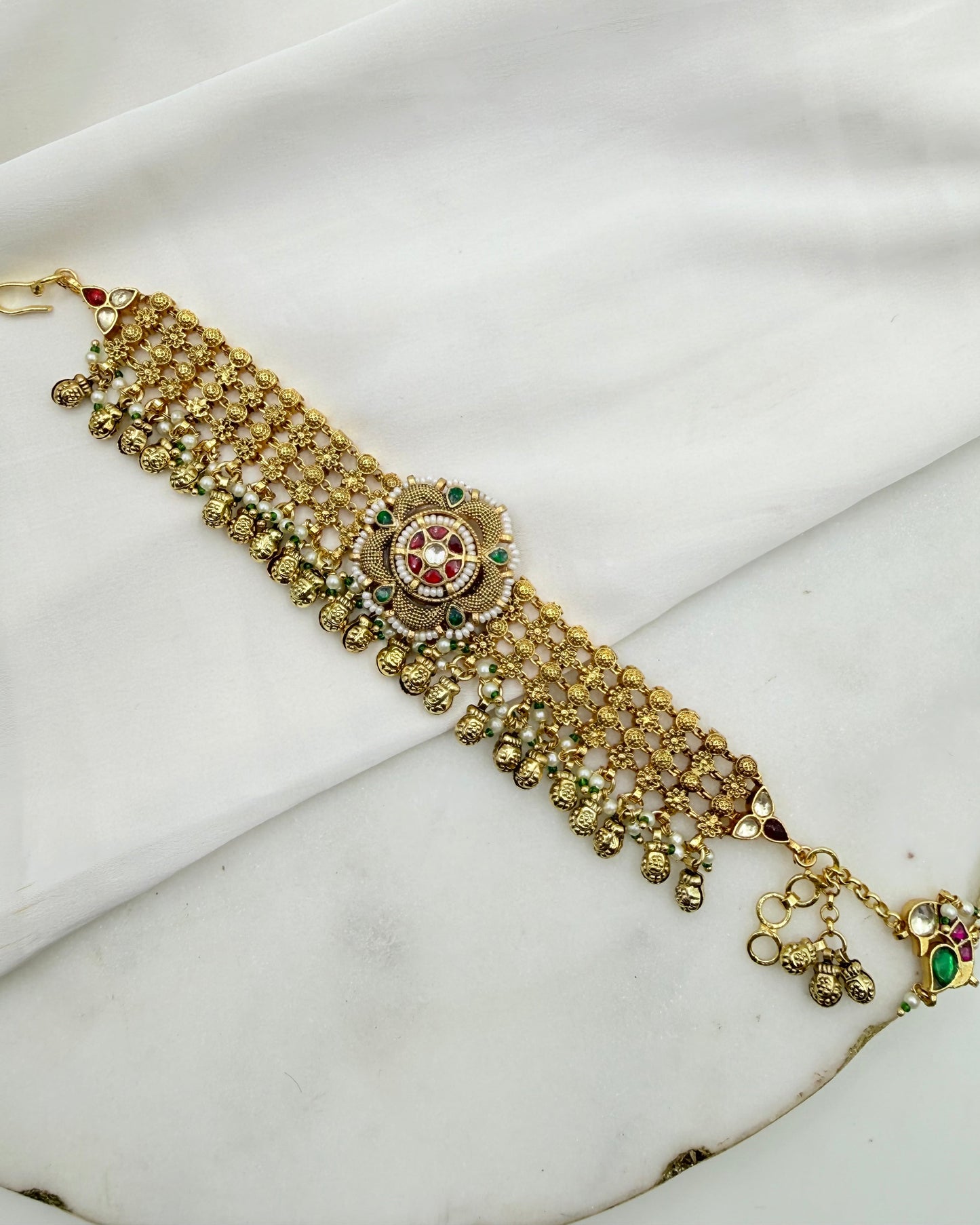 An ornate gold-toned traditional Indian choker necklace with intricate beadwork, a central floral motif with red and green stones, and dangling bell details, displayed on a white fabric background.