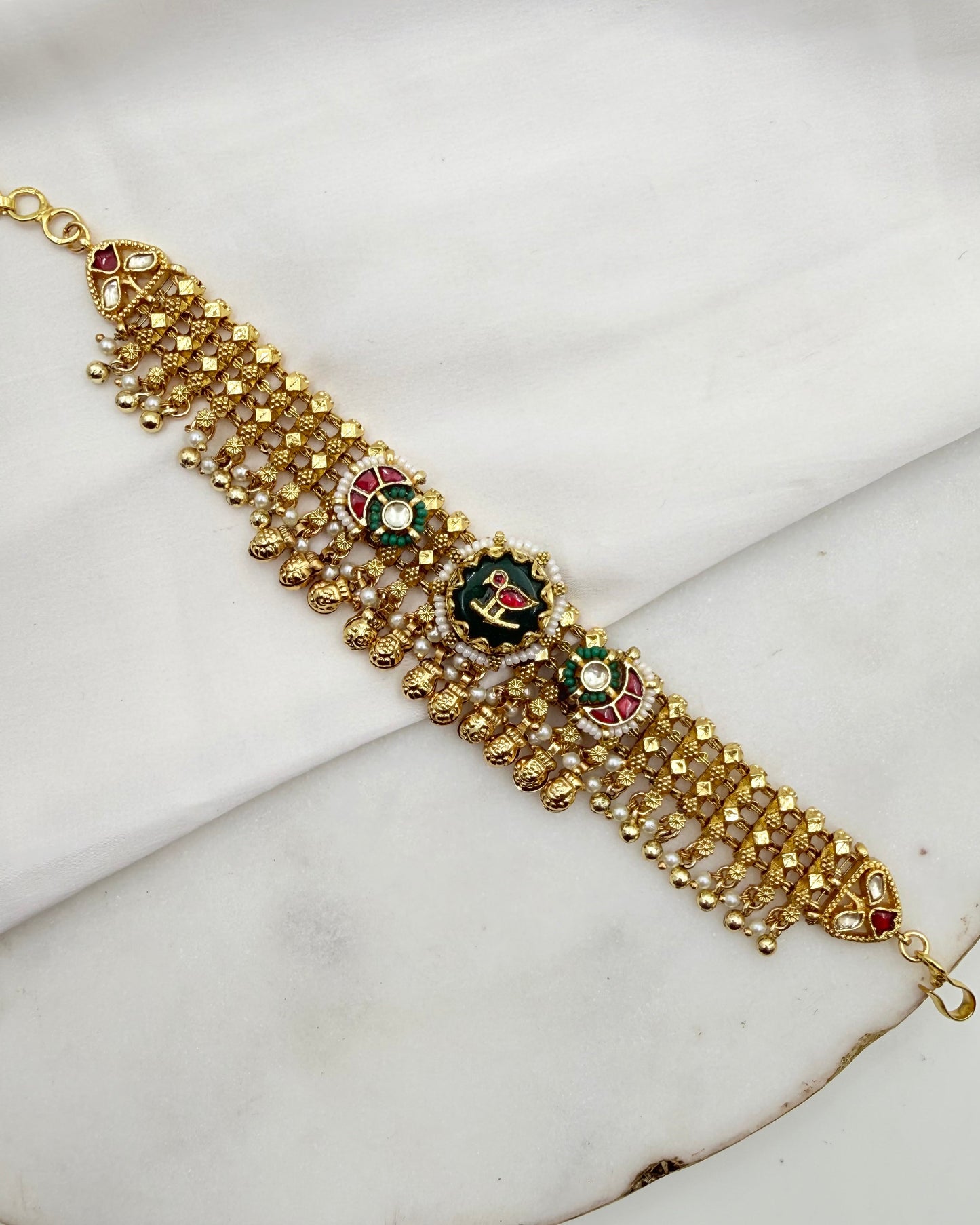 A gold-plated traditional Indian Kundan bracelet with intricate beadwork and green, red, and white enamel details, displayed on a white marble surface.