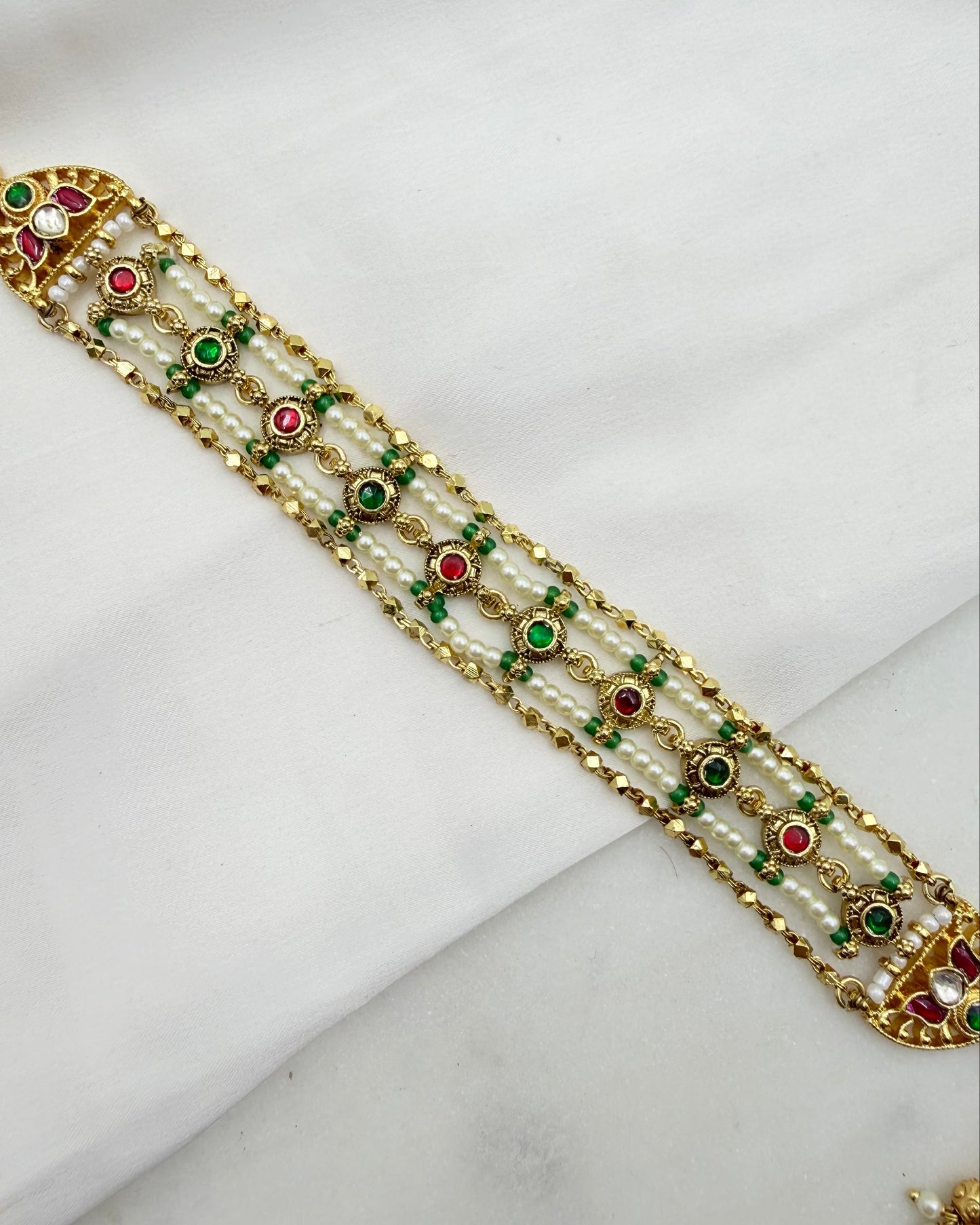 An ornate gold-plated traditional Indian bracelet decorated with white pearls, green and red stones, and small white beads, laid diagonally on a white fabric background.