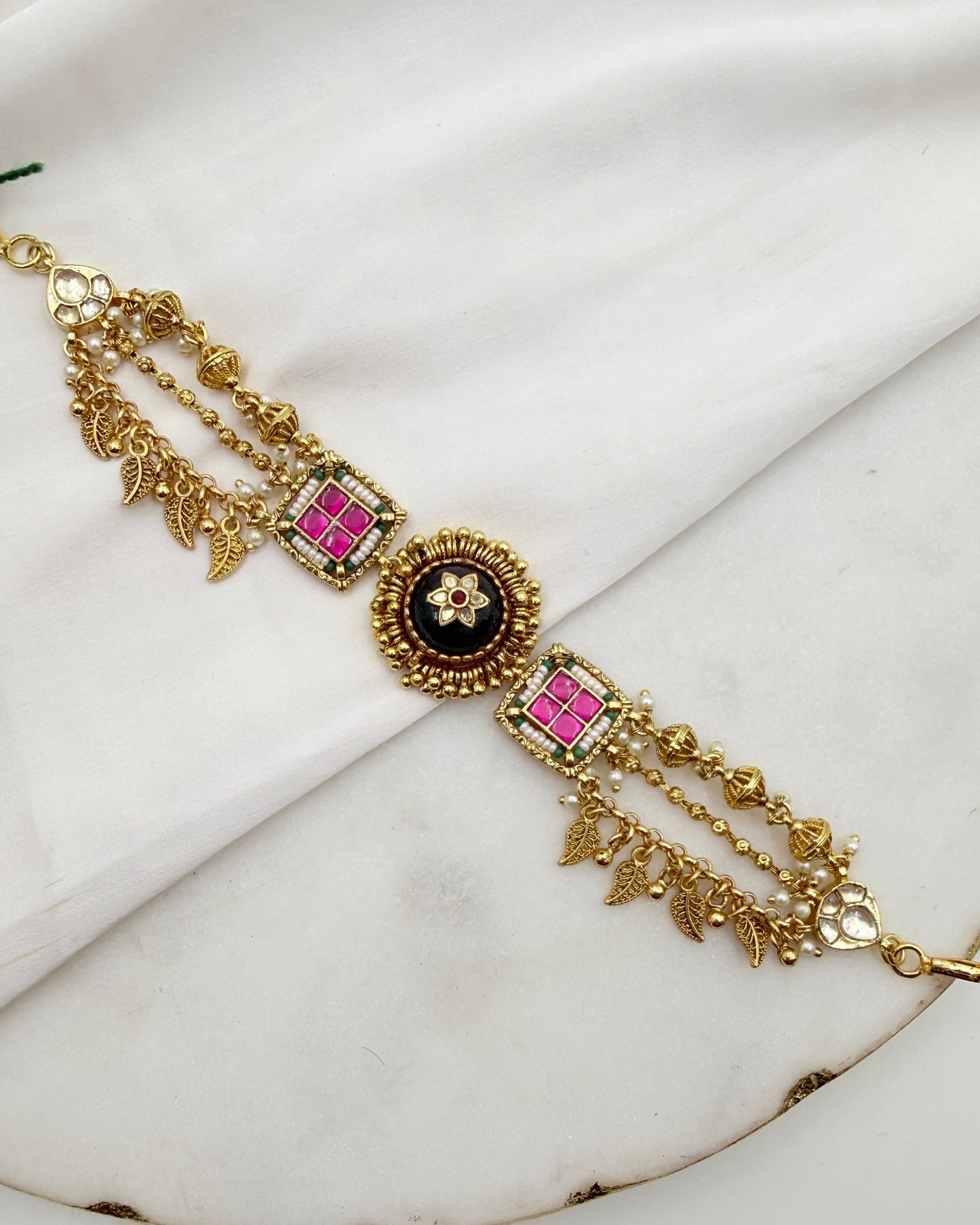 An ornate gold-toned traditional Indian bracelet with three dangling chains, a central black oval medallion with a floral motif, and two square pink enamel panels bordered with green stones, displayed on a white marble surface.