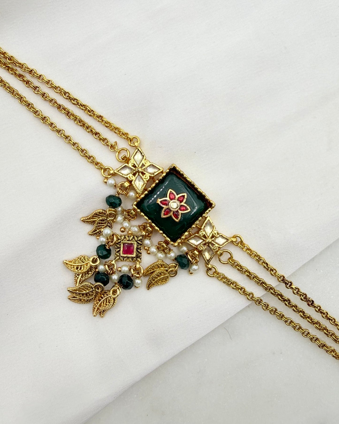 Gold-plated double-strand chain necklace with a square green enamel pendant featuring a red and white floral motif, adorned with gold leaf charms, green beads, and a small red stone, displayed on a white textured fabric.