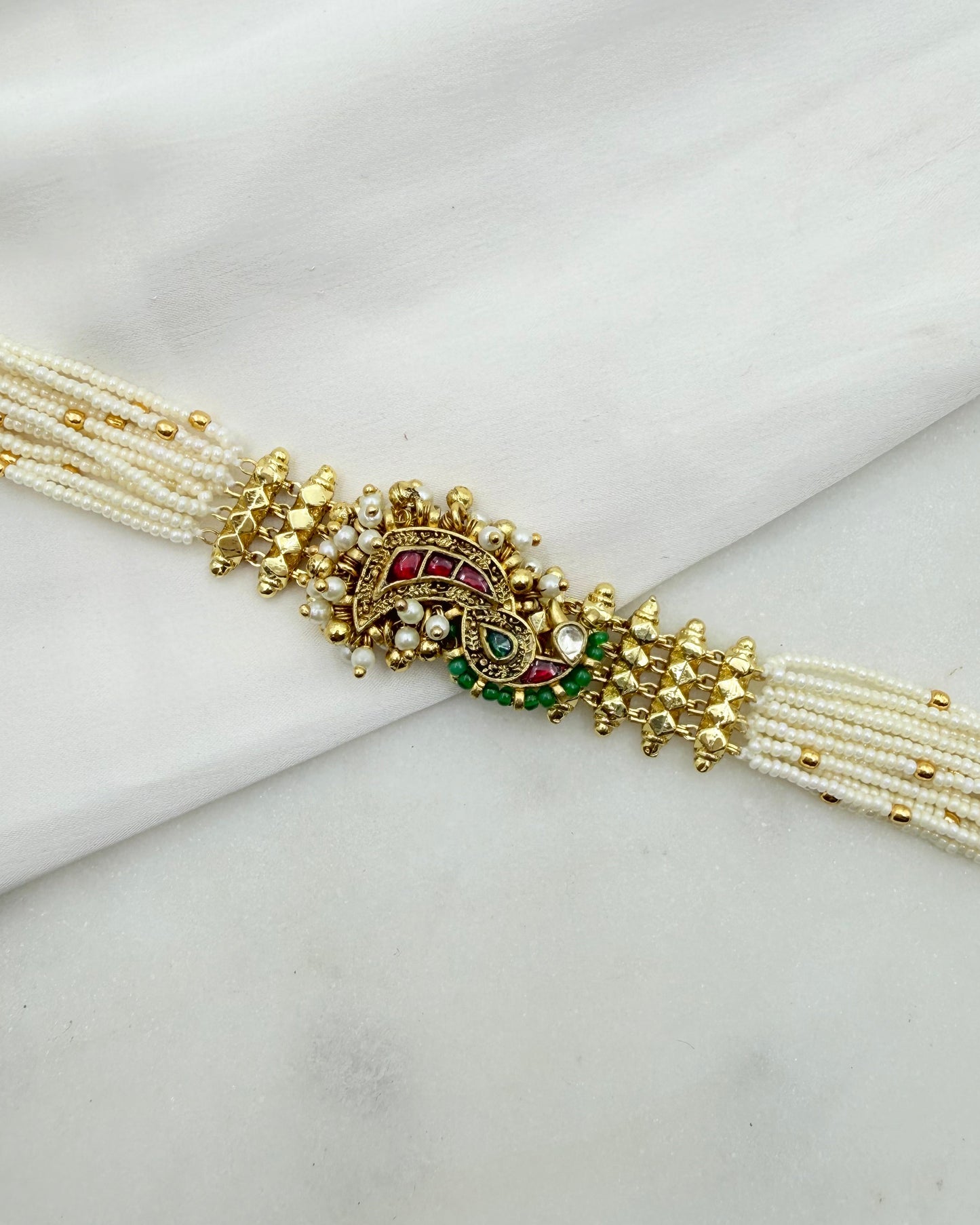 A traditional Indian pearl bracelet (sikhara) with a gold-plated peacock motif centerpiece, decorated with red and green stones and white faux pearls, displayed on a white fabric background.