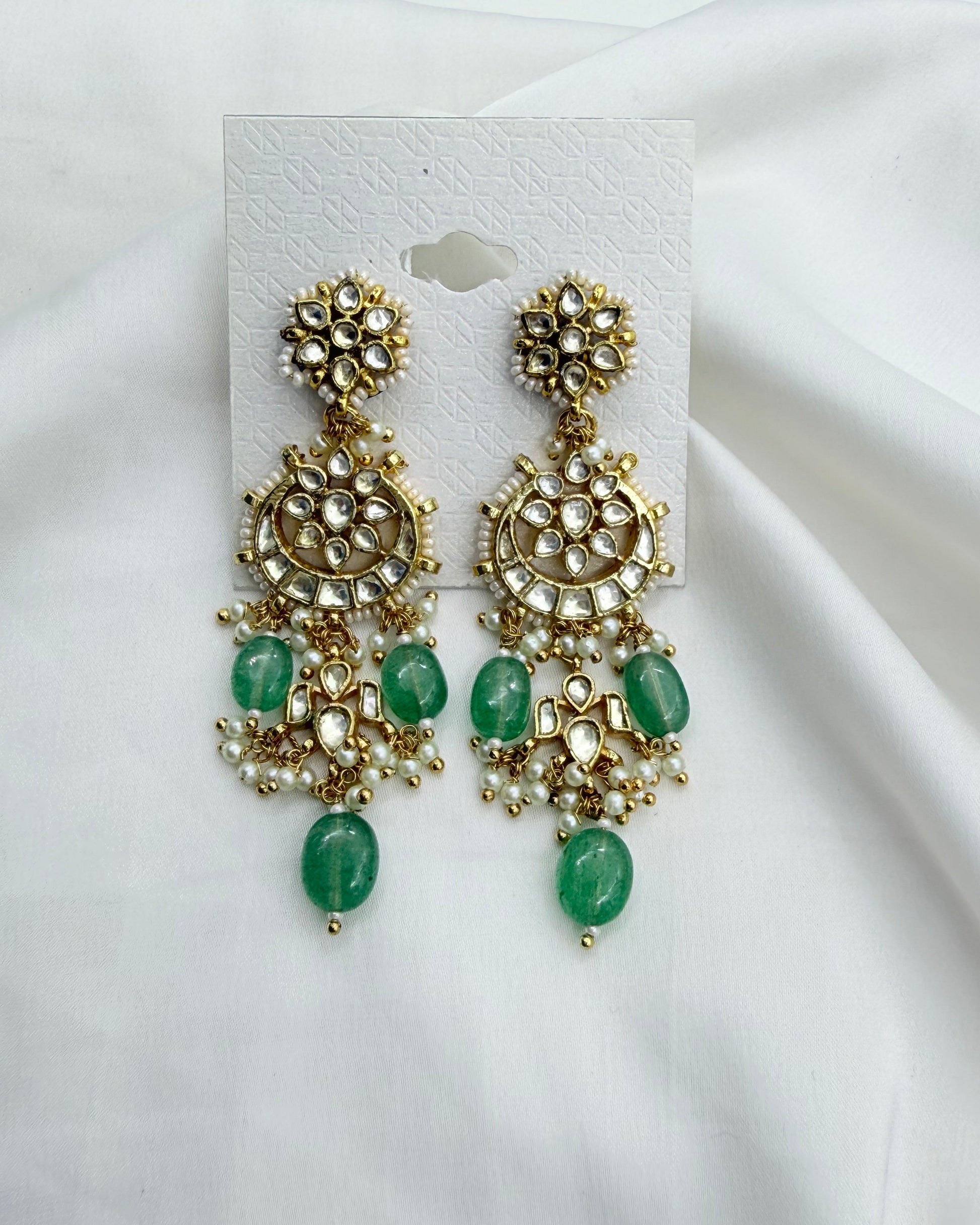 A pair of gold-toned chandelier earrings with green stone accents, displayed on a white card against a draped white fabric background.