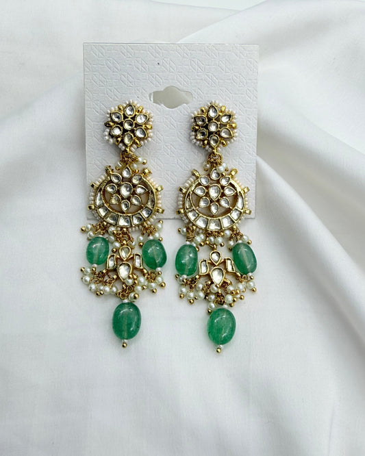 A pair of gold-toned chandelier earrings with green stone accents, displayed on a white card against a draped white fabric background.
