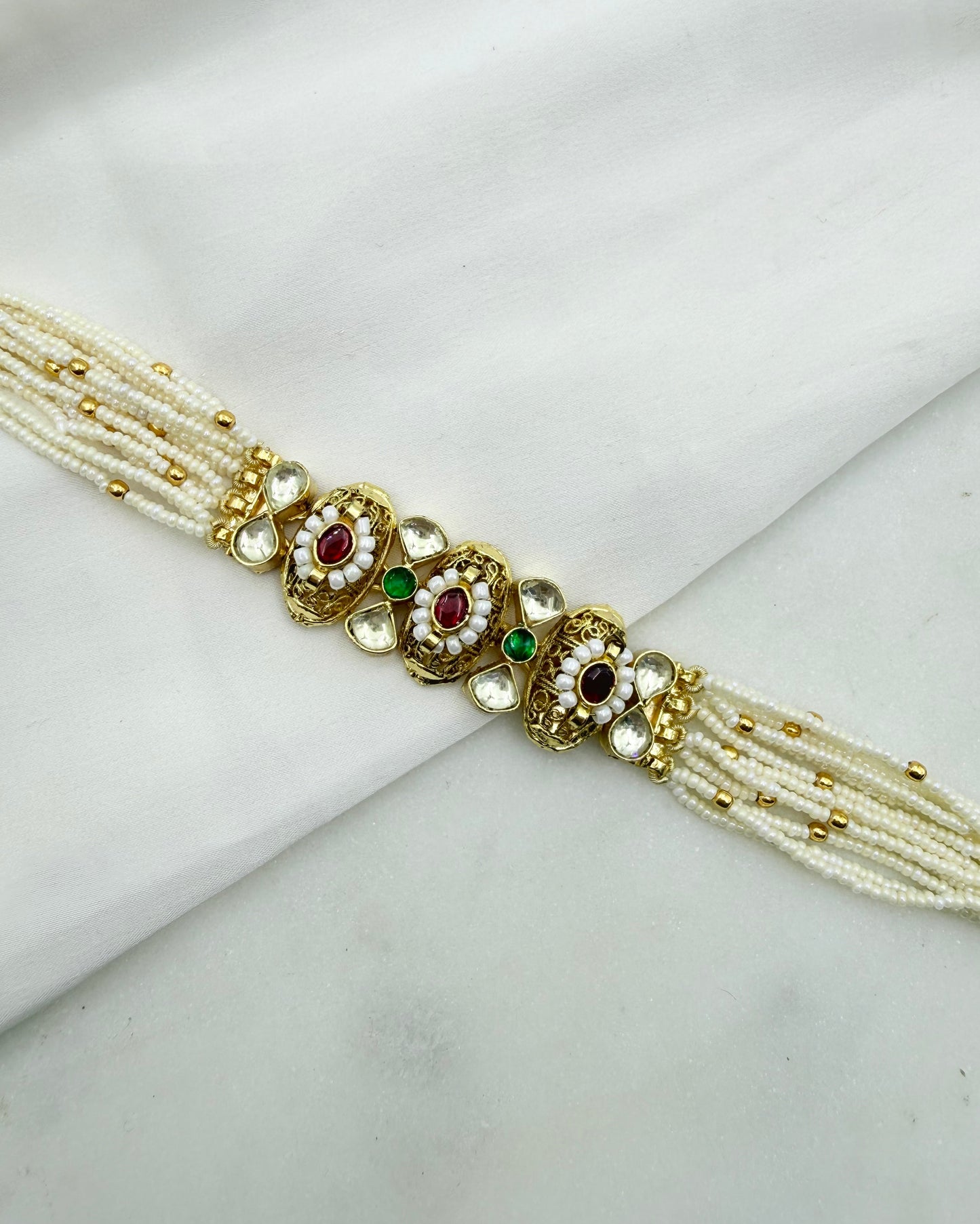 A traditional Indian pearl necklace with gold-plated borders and three ornate central floral motifs decorated with red, green, and black stones, displayed on a white fabric background.