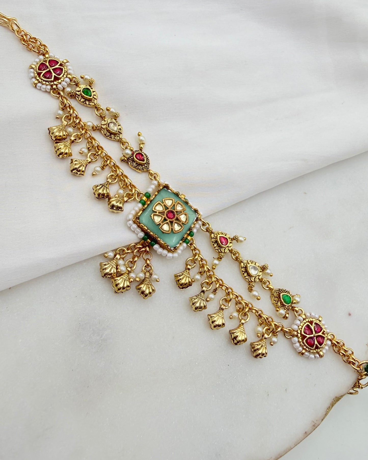 An ornate gold-toned traditional necklace with a central turquoise-green square pendant, decorated with red and green stone accents, white pearl-like beads, and dangling floral and bell-shaped charms, displayed on a white fabric background.