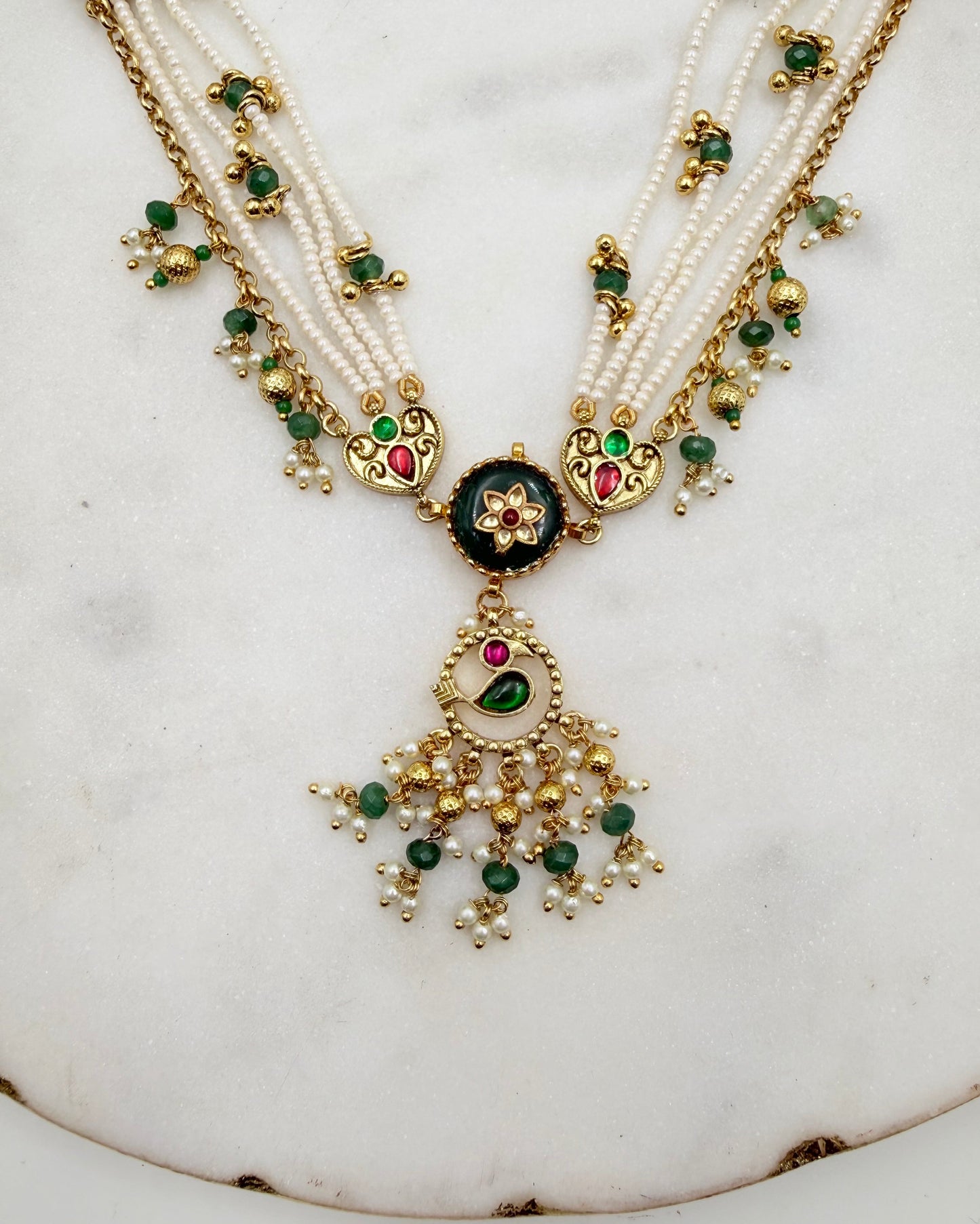 An ornate traditional Indian necklace featuring multiple strands of white pearls, gold-plated chains, and a central black enamel medallion with green and red stones, hanging from a large gold tassel decorated with green and white pearls.