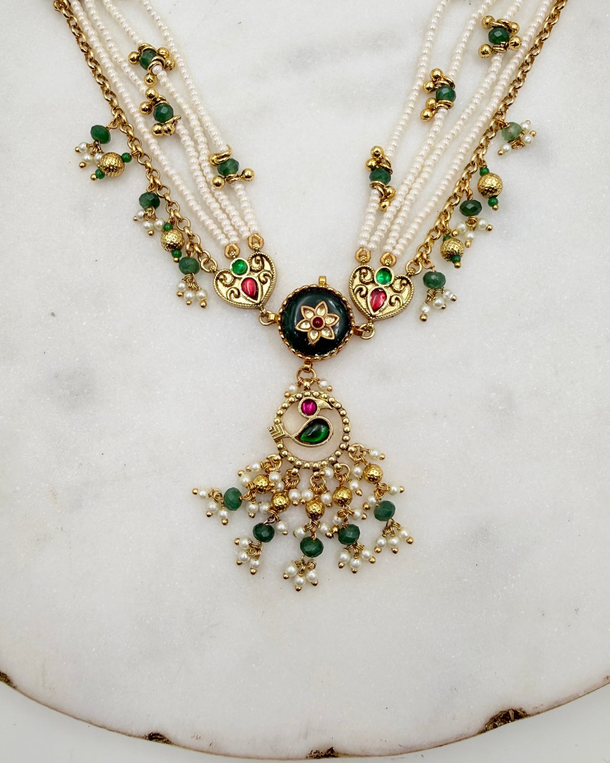 An ornate traditional Indian necklace featuring multiple strands of white pearls, gold-plated chains, and a central black enamel medallion with green and red stones, hanging from a large gold tassel decorated with green and white pearls.