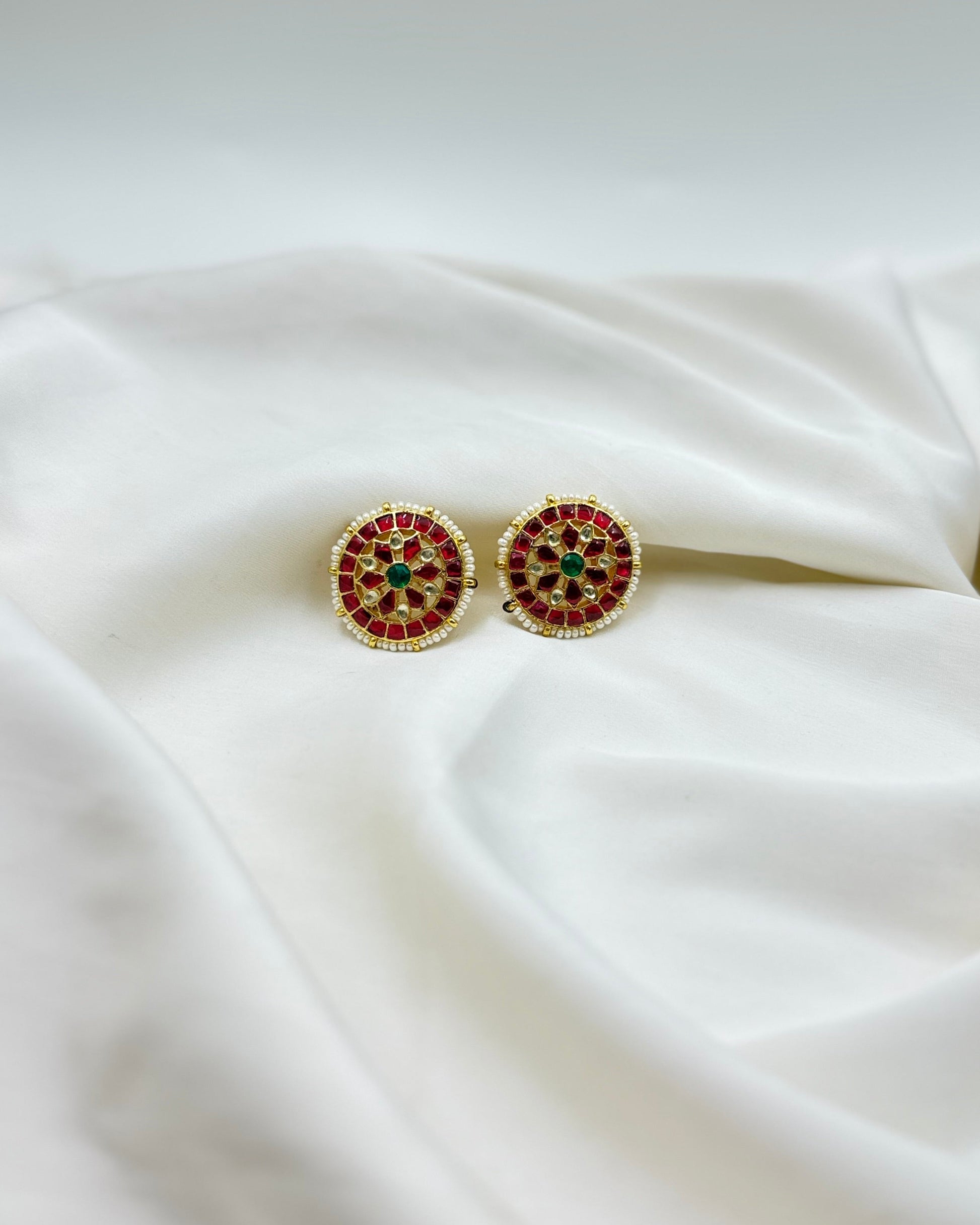 A pair of round gold-toned stud earrings with red and green stone detailing, displayed on draped white fabric.