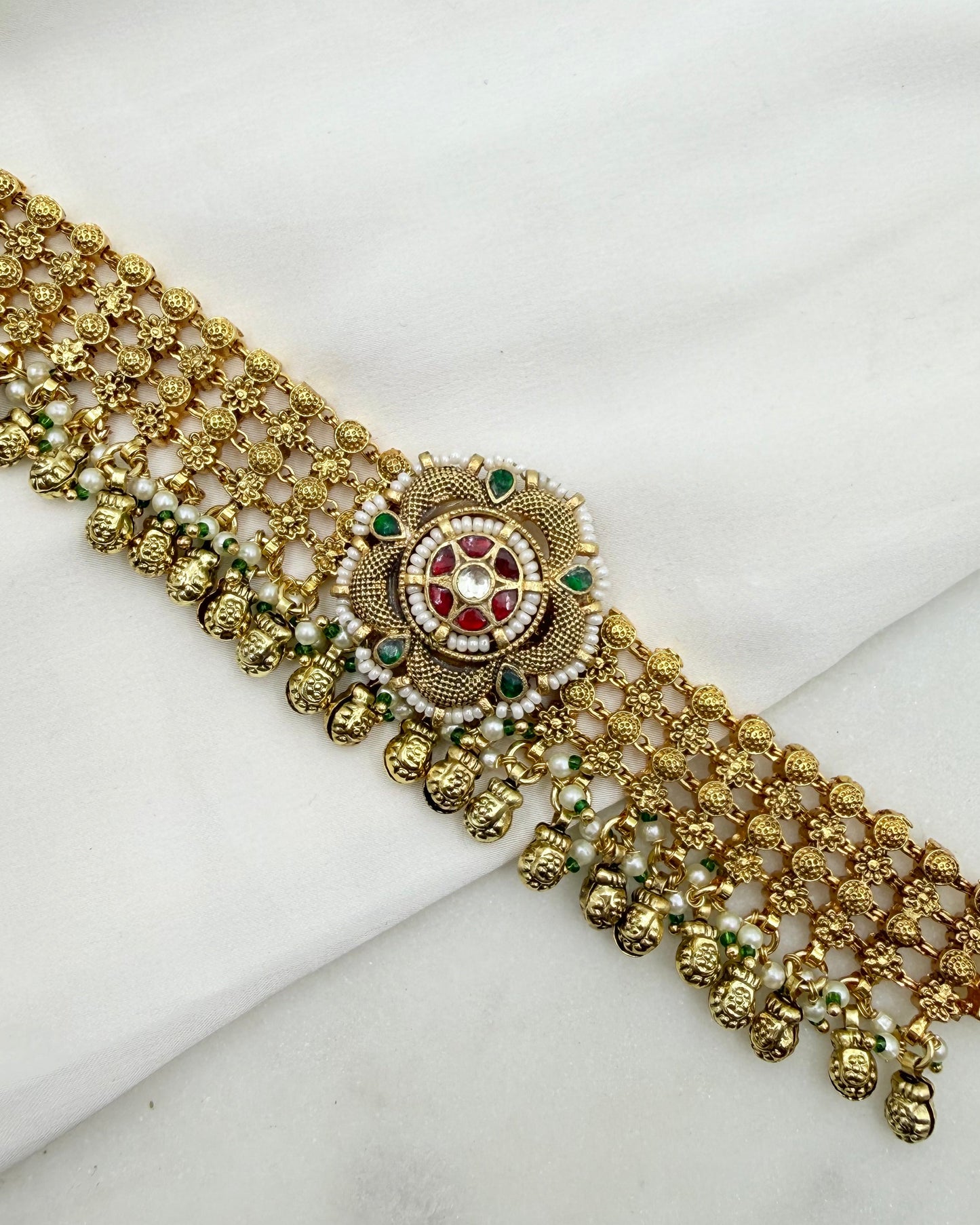 An ornate gold-toned traditional Indian choker necklace with intricate beadwork, small green and red stones, and white faux pearls, displayed on a white fabric background.