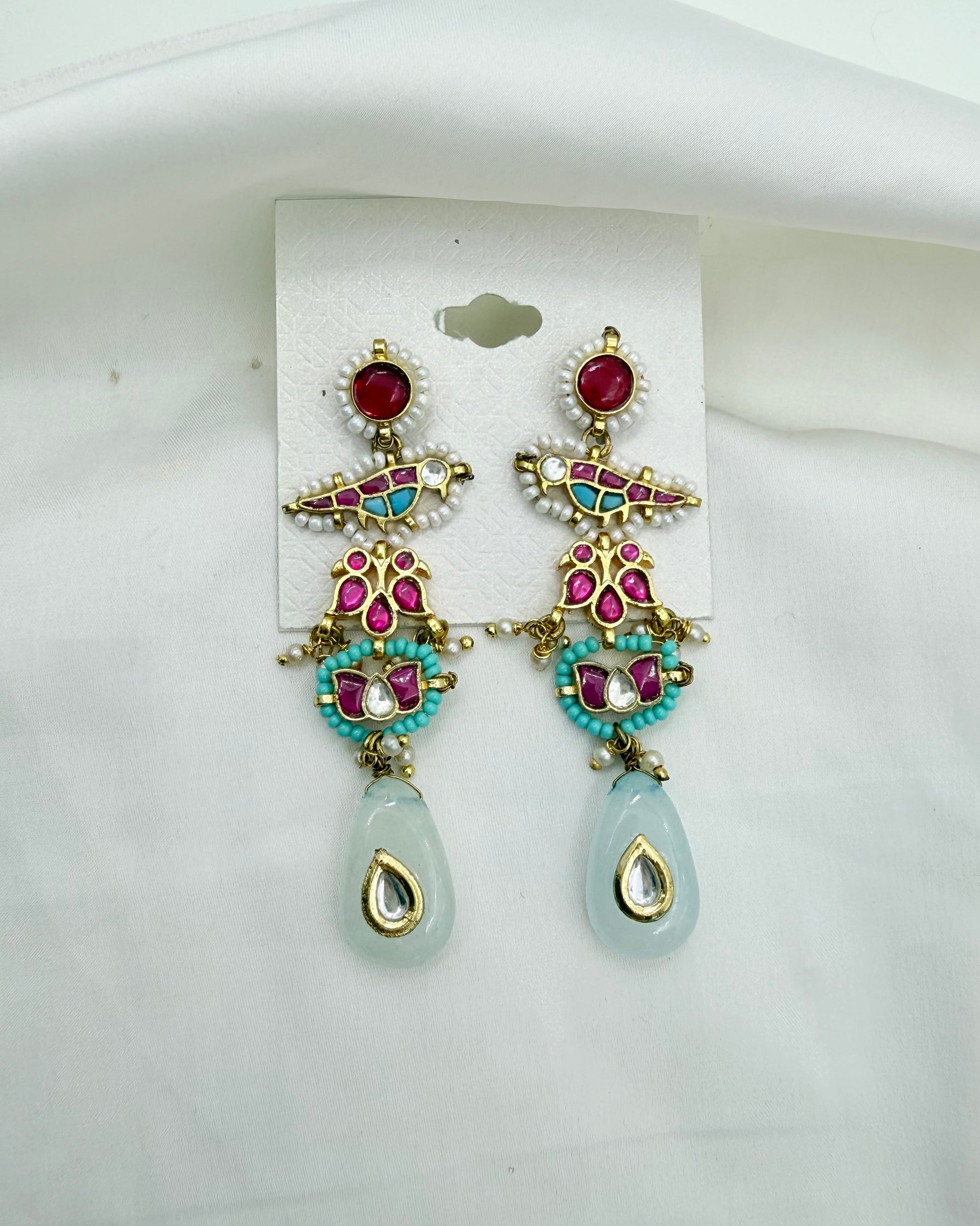 A pair of ornate gold-plated Indian-style dangle earrings with red, turquoise, and white stones, hanging from a white display card on a white fabric background.