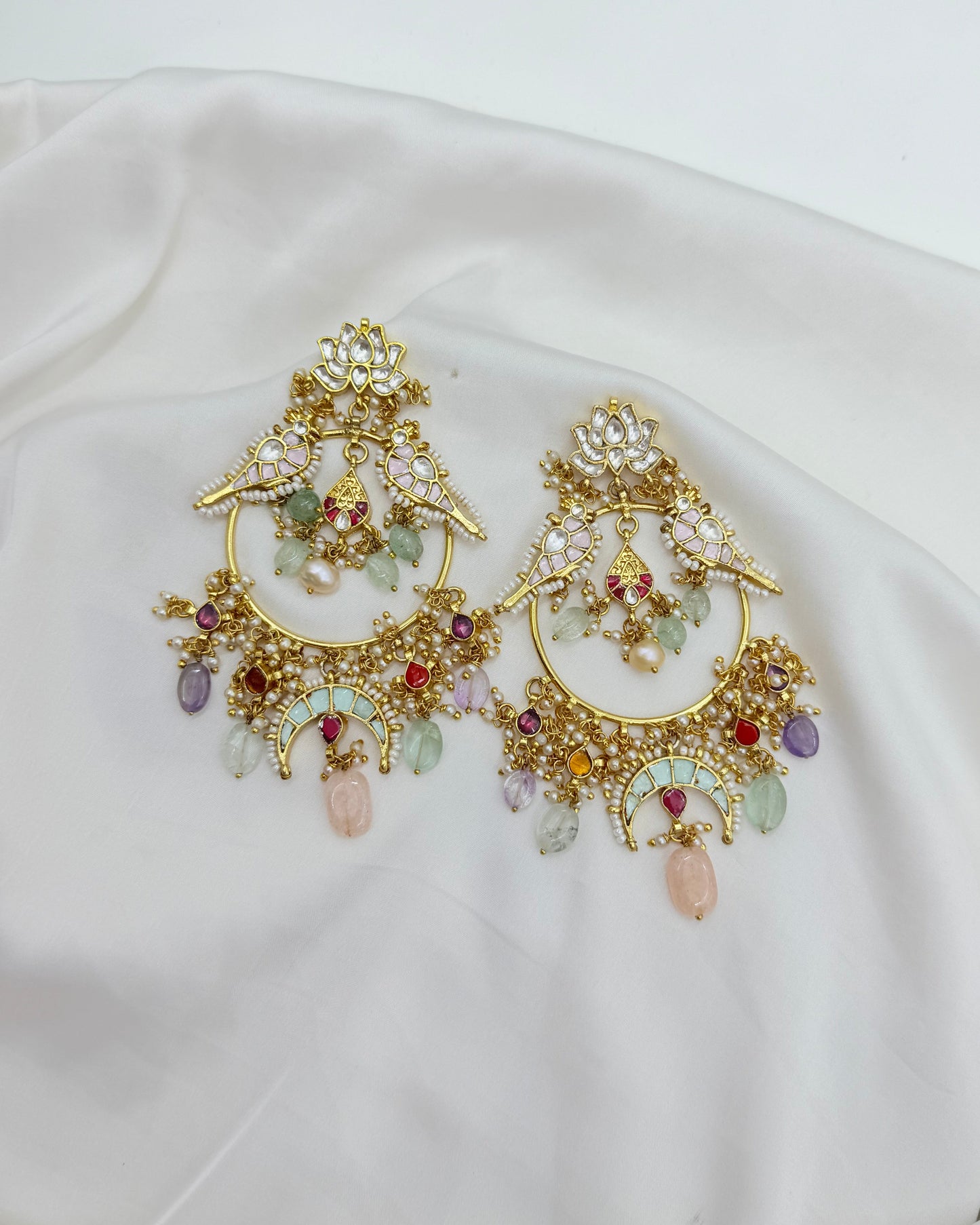 A pair of ornate gold-toned statement earrings with intricate crystal and gemstone detailing, displayed on a draped white satin fabric.