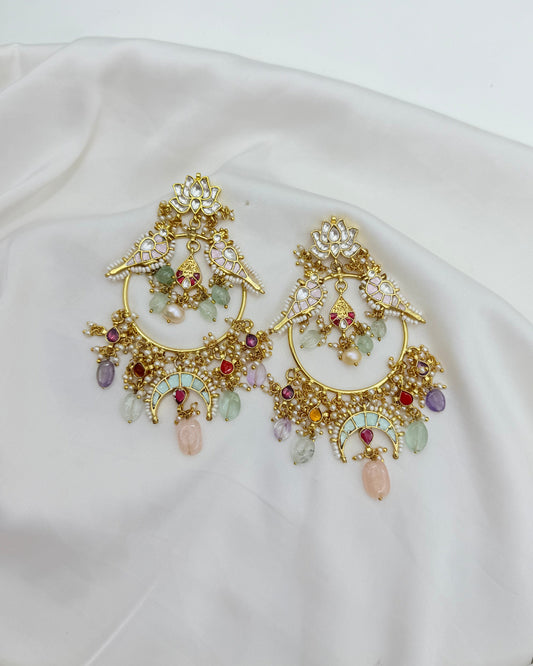 A pair of ornate gold-toned statement earrings with intricate crystal and gemstone detailing, displayed on a draped white satin fabric.